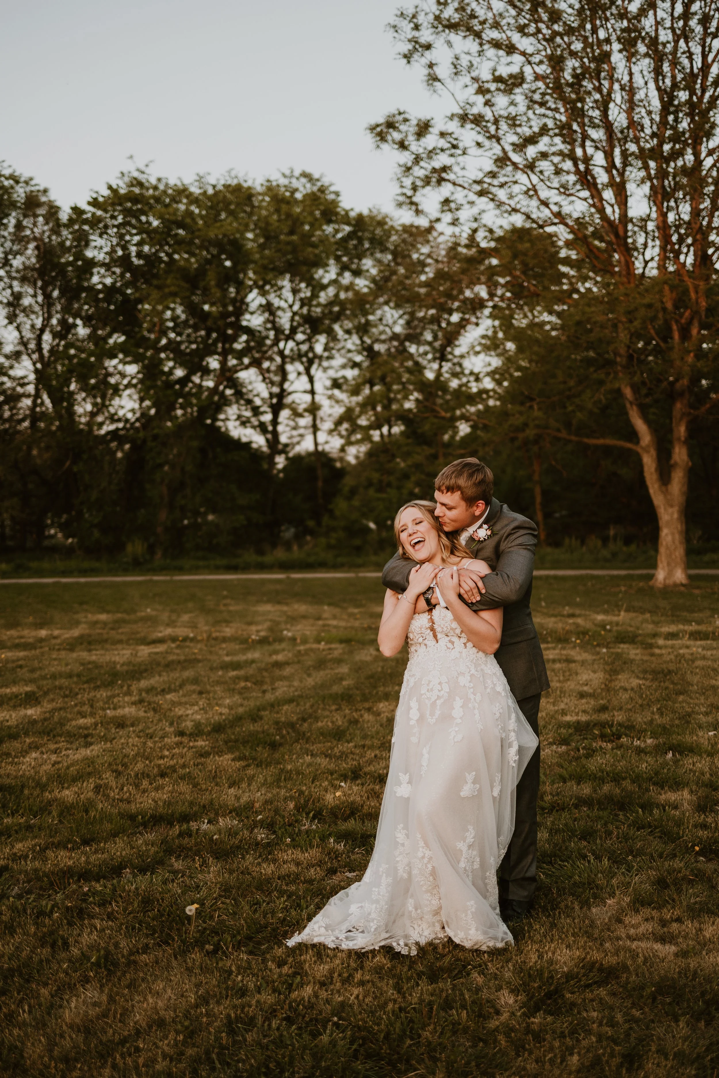 A happy couple in wedding attire embracing outdoors at sunset, with trees in the background.