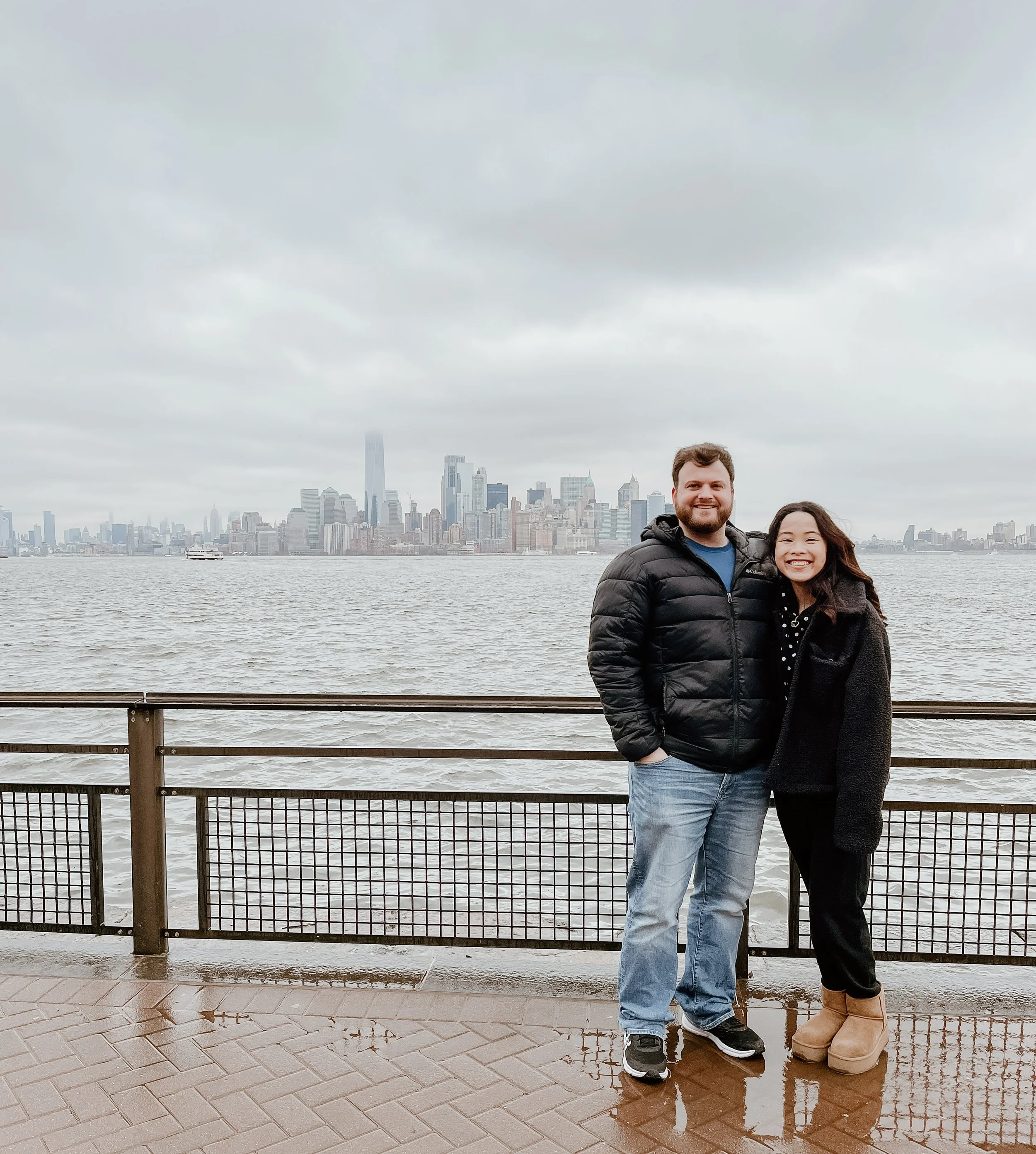 A man and woman standing together by a waterfront with a city skyline in the background on a cloudy day.
