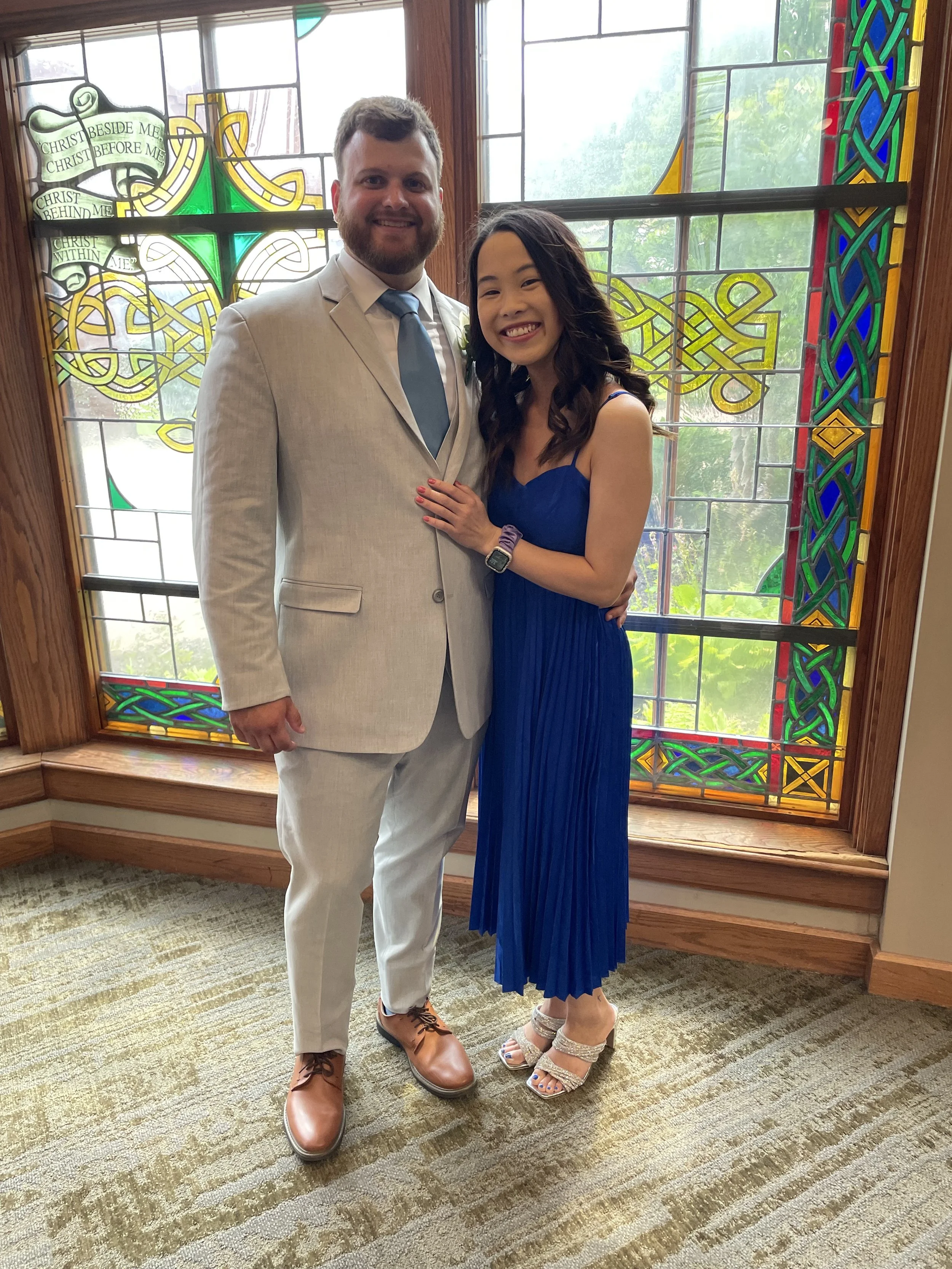 A man in a light gray suit and a woman in a blue dress standing together, smiling, in front of a stained glass window with colorful designs and green foliage outside.