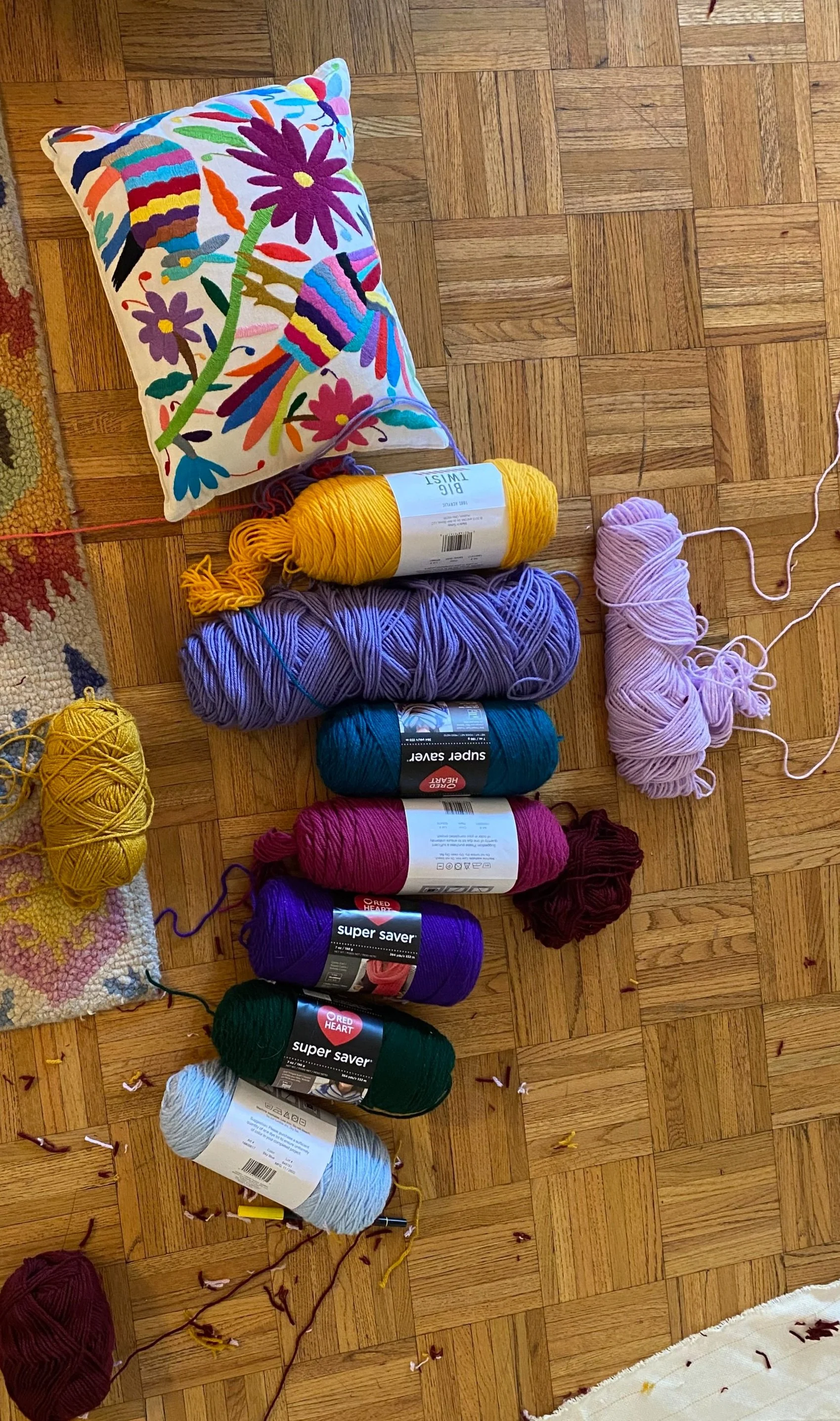 Colorful embroidered pillow, several skeins of yarn in various colors, and a small portion of a braided carpet on a wooden floor.