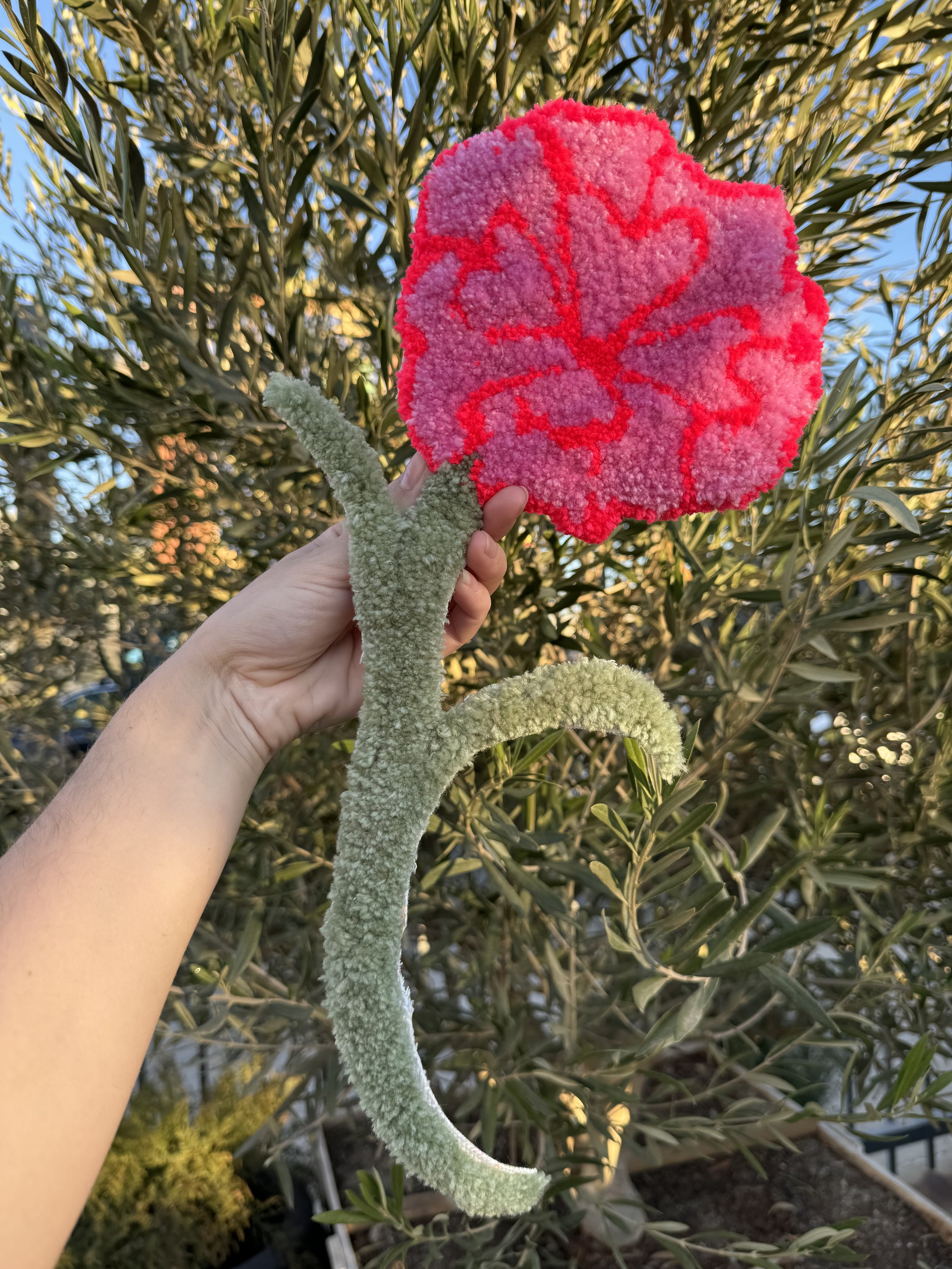 A person holding a colorful, handmade, flower-shaped craft with a fuzzy green stem and a pink and red petals, set against a background of leafy bushes and a clear blue sky.