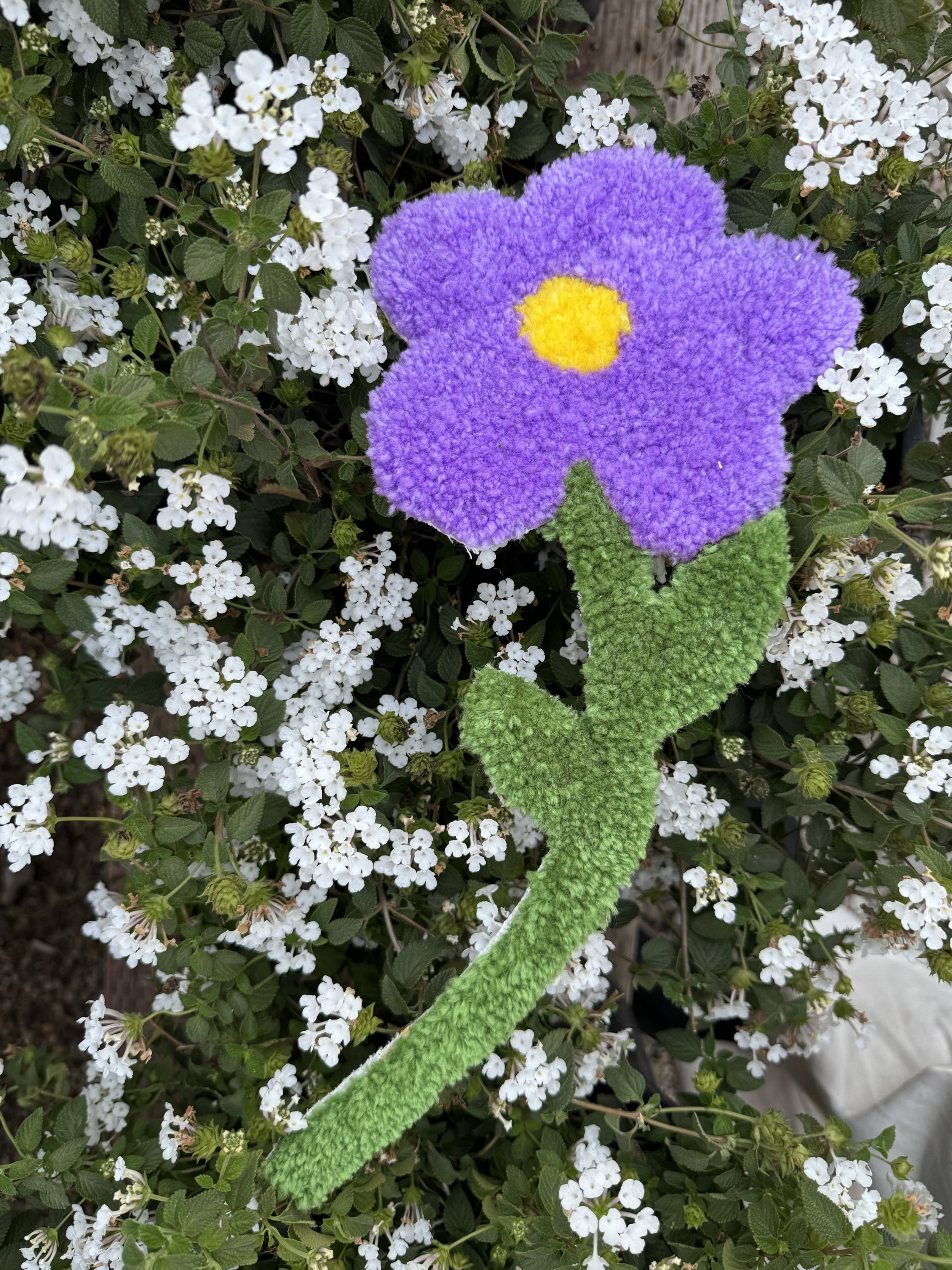 A purple flower with a yellow center and green stem, made from craft materials, laid on a background of small white flowers and green leaves.