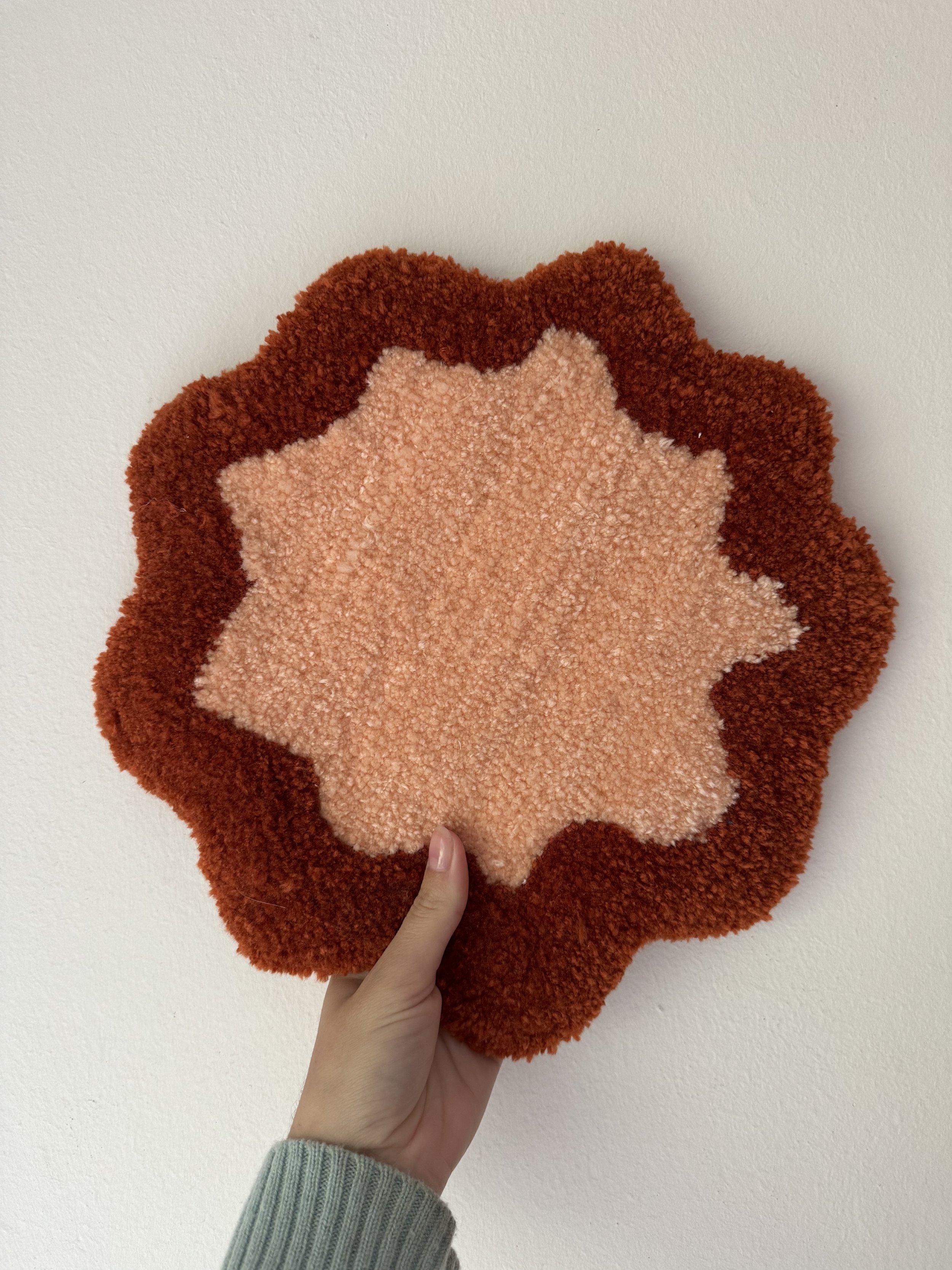 Person holding a plush, star-shaped rug with a pink center and a dark red and rust-colored edge, against a plain light-colored wall.