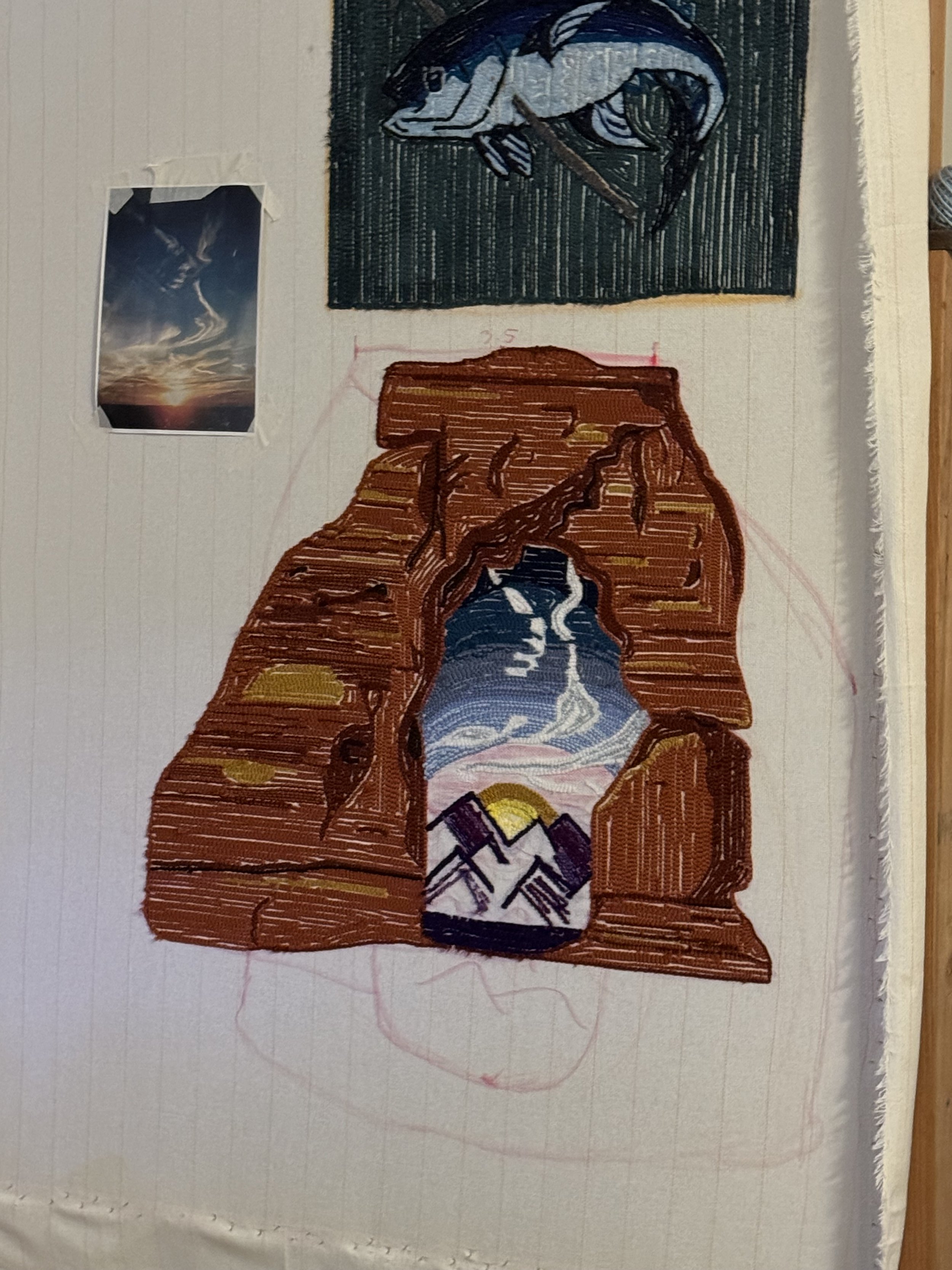 A partially completed embroidery of a mountain scene inside a canyon with a river, on brown rock formations, on a piece of woven fabric. A small reference photo of a sunset over clouds is taped to the fabric.