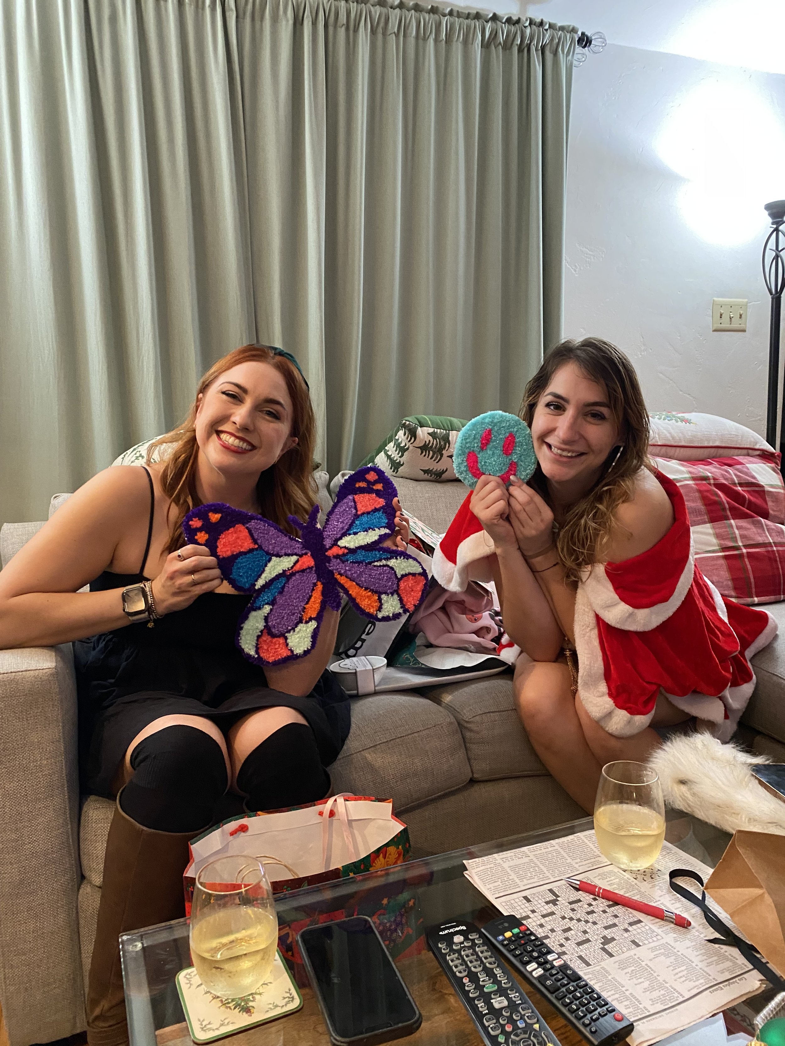 Two women sitting on a couch, smiling and holding colorful knitted crafts, one butterfly and one smiley face, in a living room decorated for Christmas.