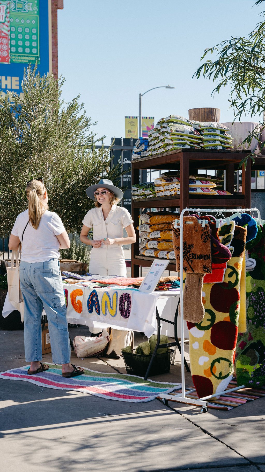 Two women talking at an outdoor craft market stall selling colorful woven rugs and wall hangings on a sunny day.