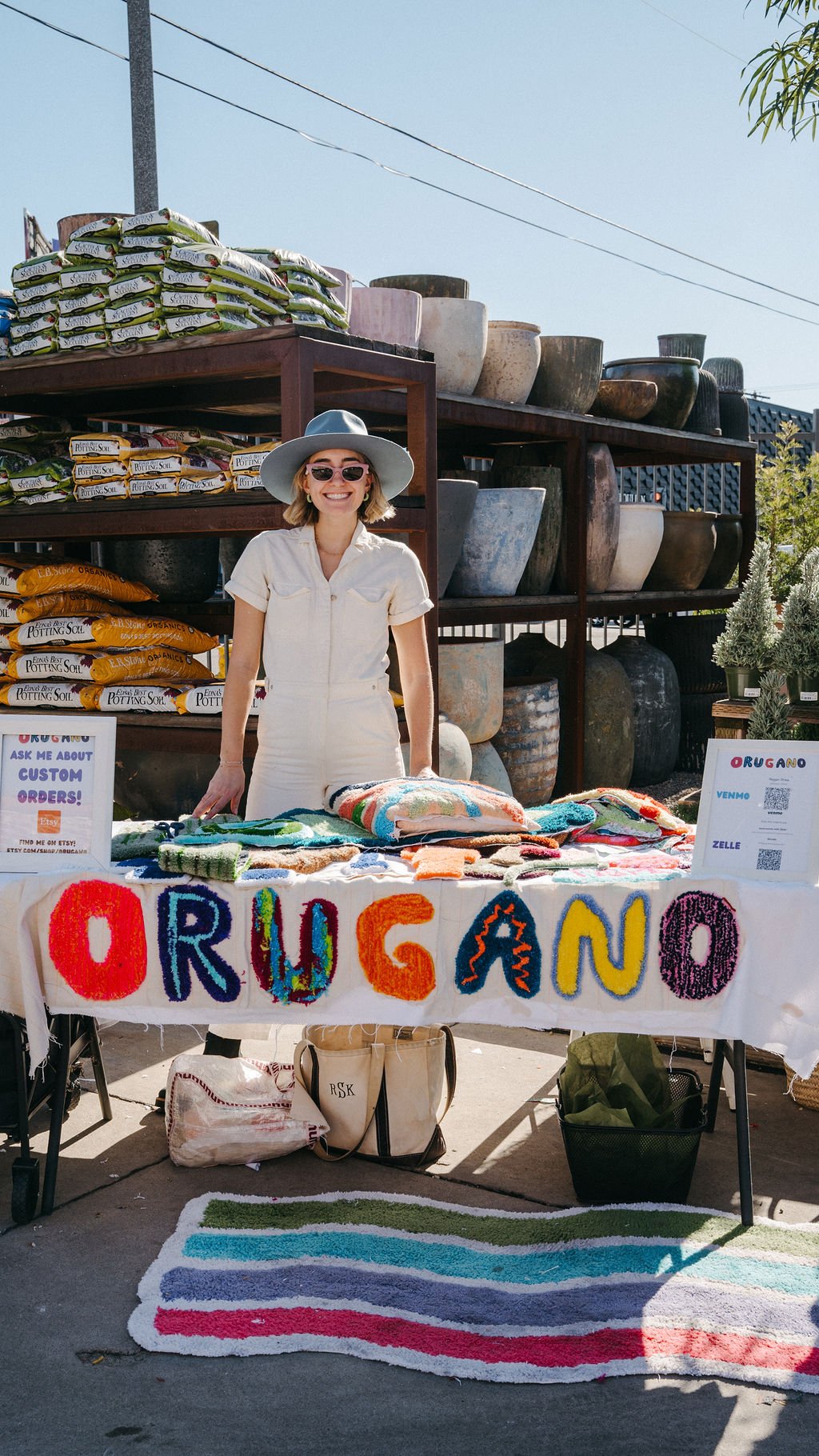 A woman selling colorful handmade yarn items at an outdoor market stall with a sign reading 'ORUGANO'. She is wearing a light-colored dress and a wide-brimmed hat, smiling, with various bags and a rainbow-striped rug on the ground in front of her.