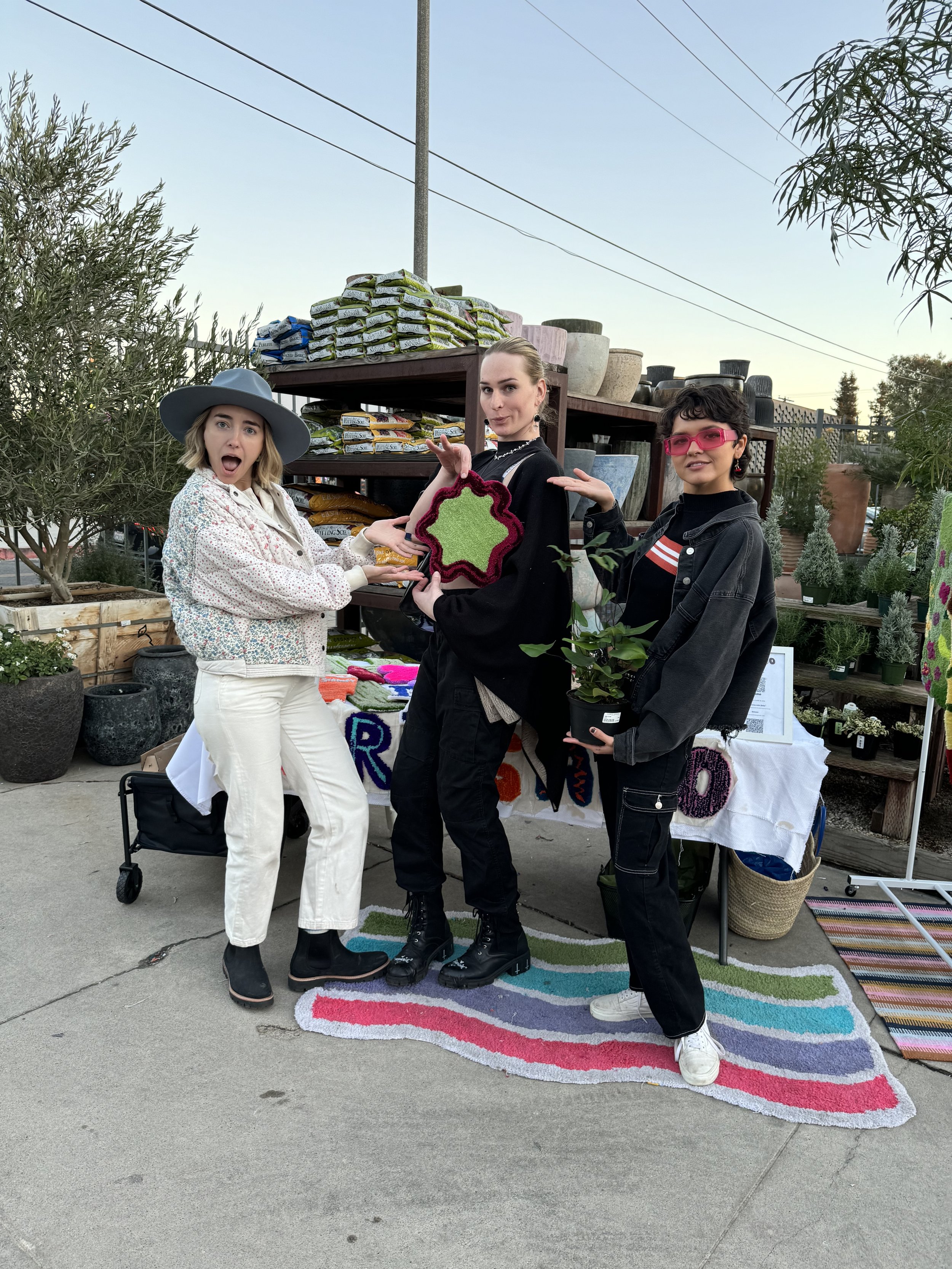 Three women standing at a sidewalk stall selling colorful handmade crafts, with plants and gardening supplies behind them.