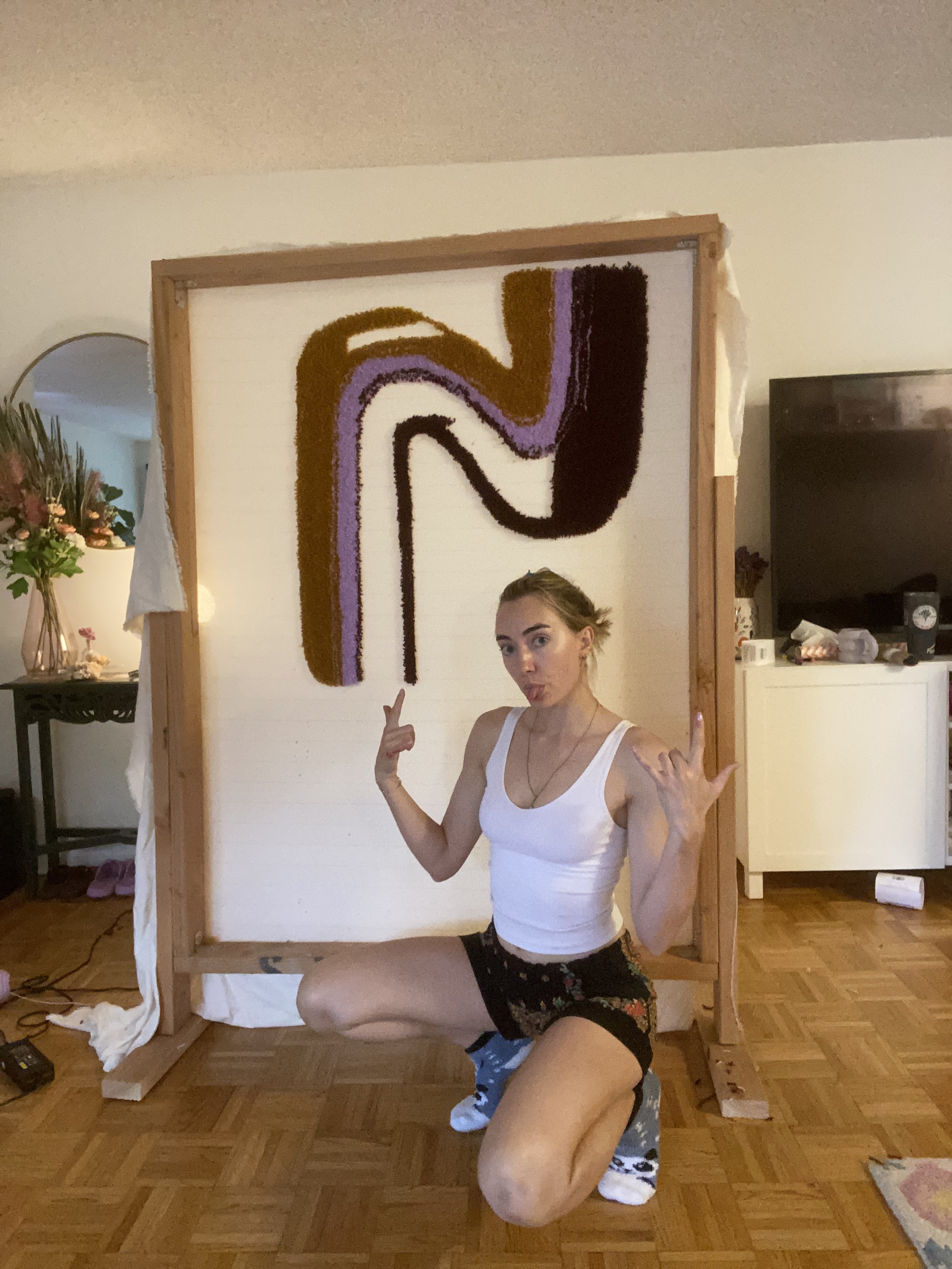 Young woman in white tank top and floral shorts posing in front of a large, colorful, textured artwork with her hands gesturing upward.