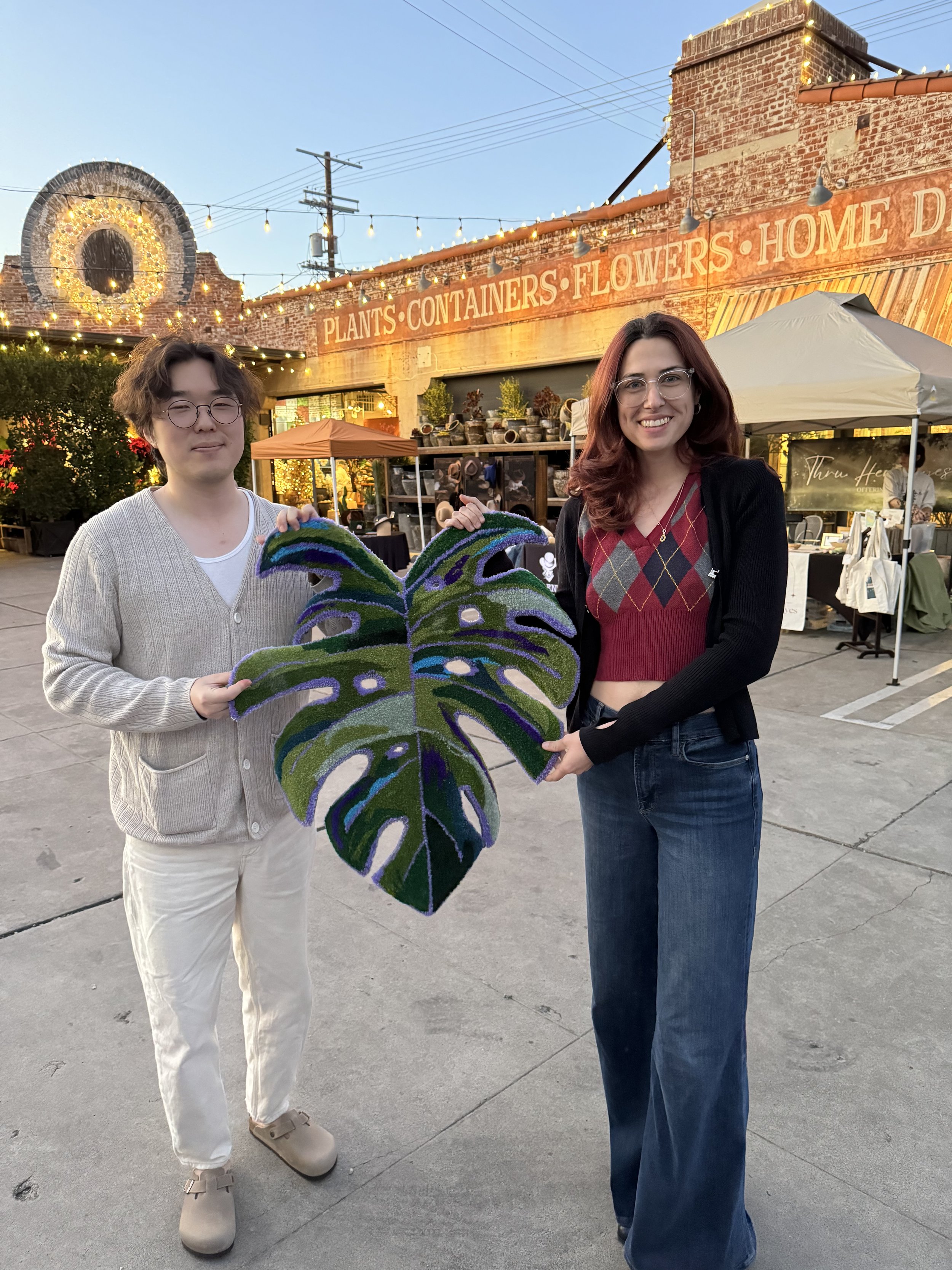 Two people holding a large green and purple monstera leaf decoration in an outdoor market or shopping area at dusk with string lights, brick building, and vendors in the background.