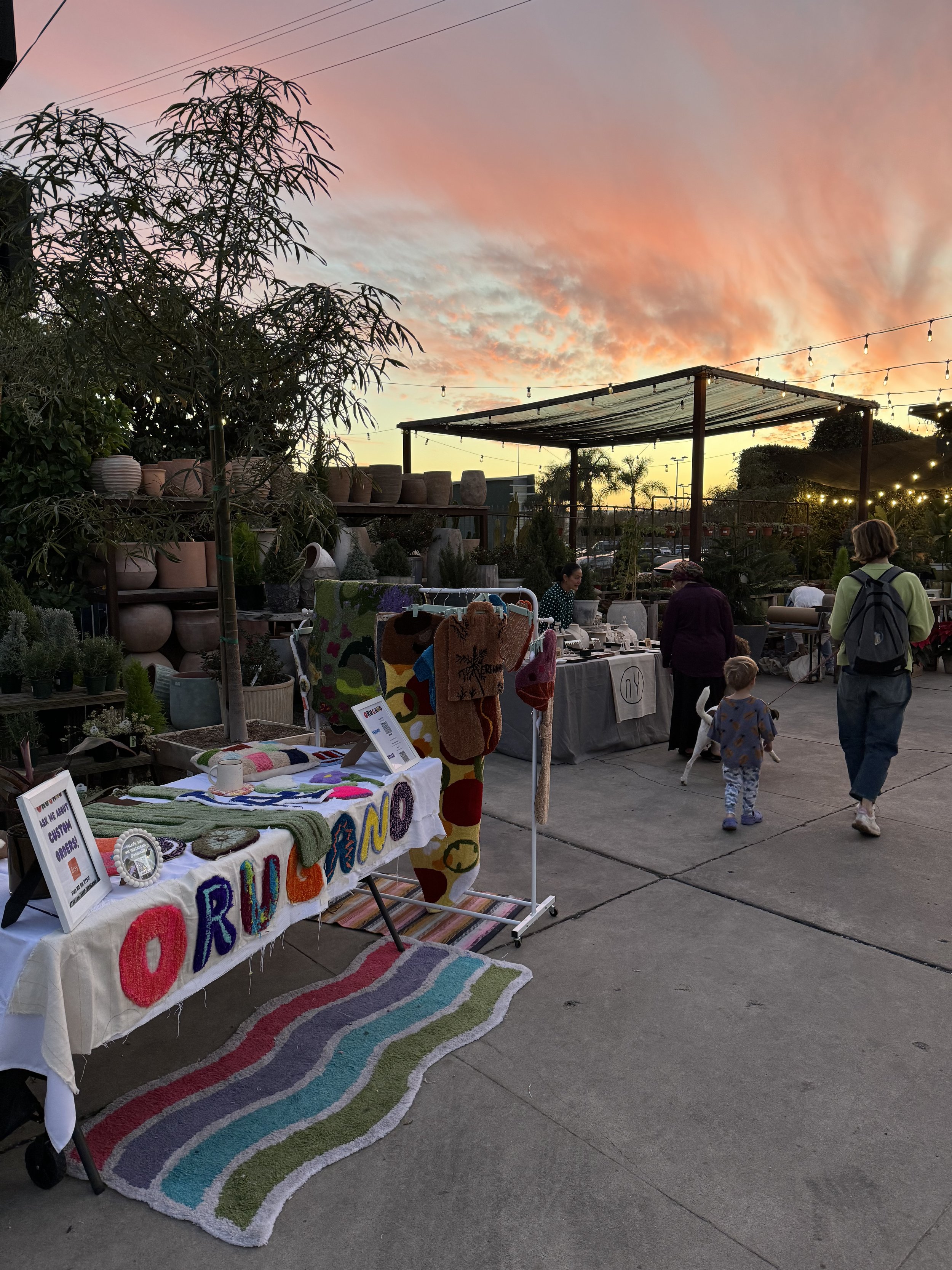Outdoor market setup at sunset with tables selling handmade items, colorful rugs, and potted plants, with people browsing and children walking nearby.