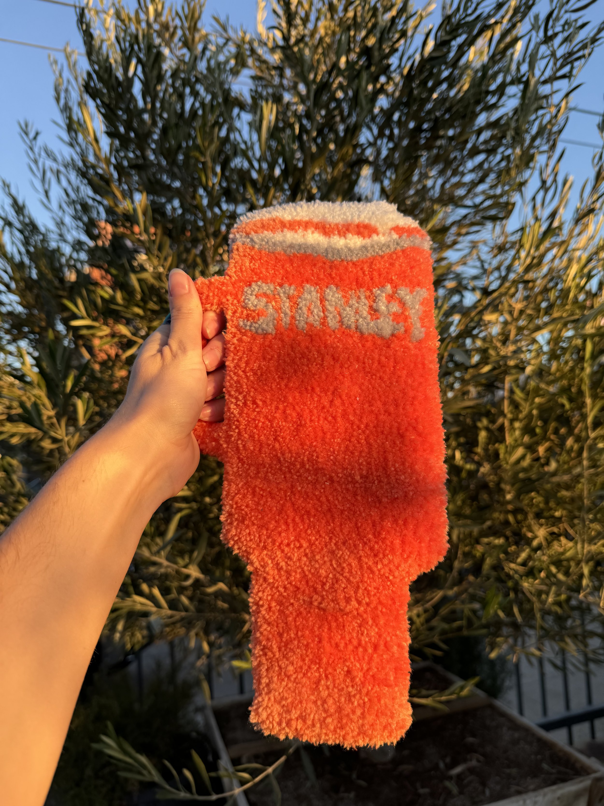 A hand holds the handle of a fuzzy, tufted rug shaped like a Stanley tumbler.