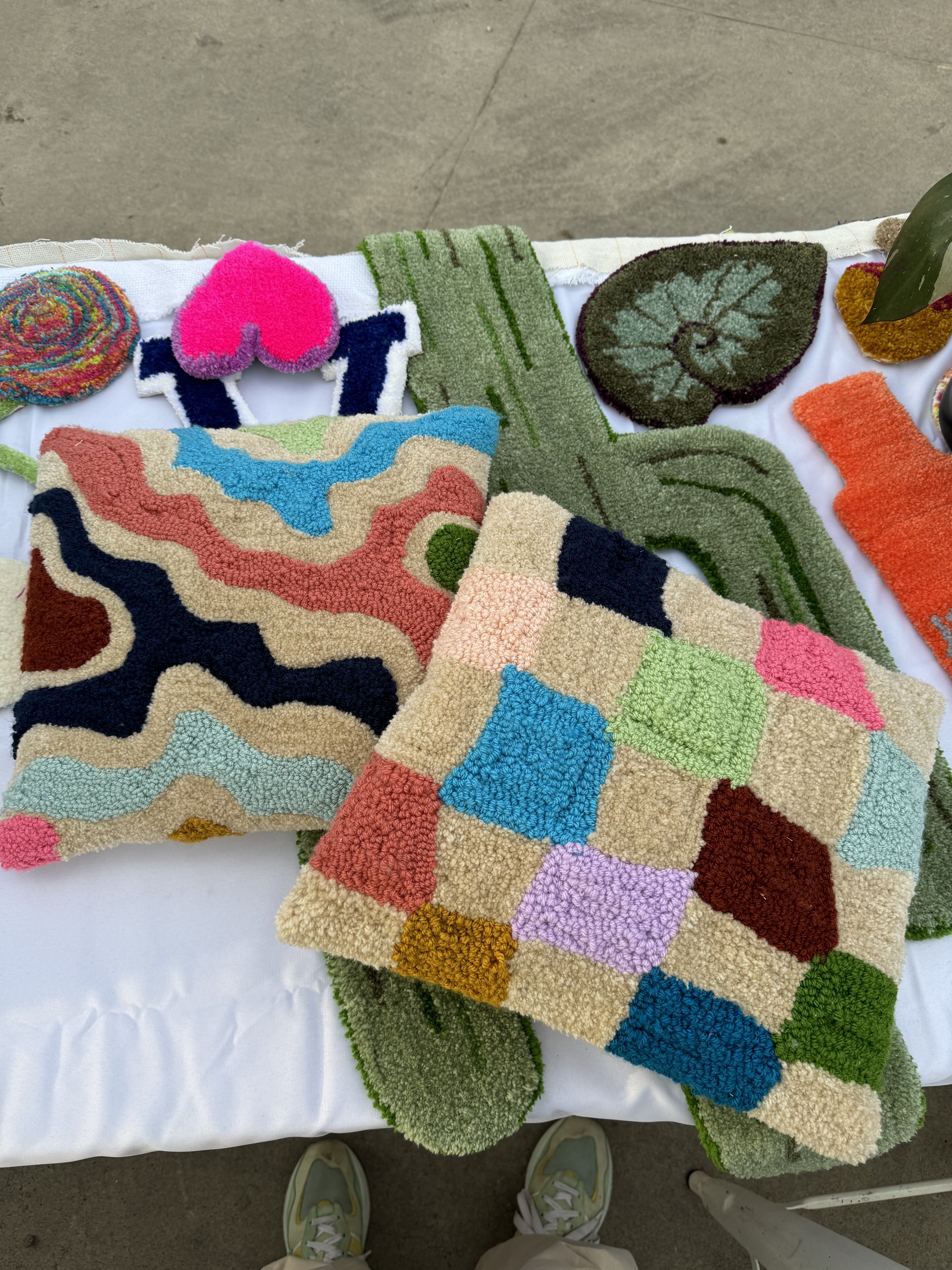 Colorful, patterned handmade rugs and pillows displayed on a white tablecloth at an outdoor market or craft fair.