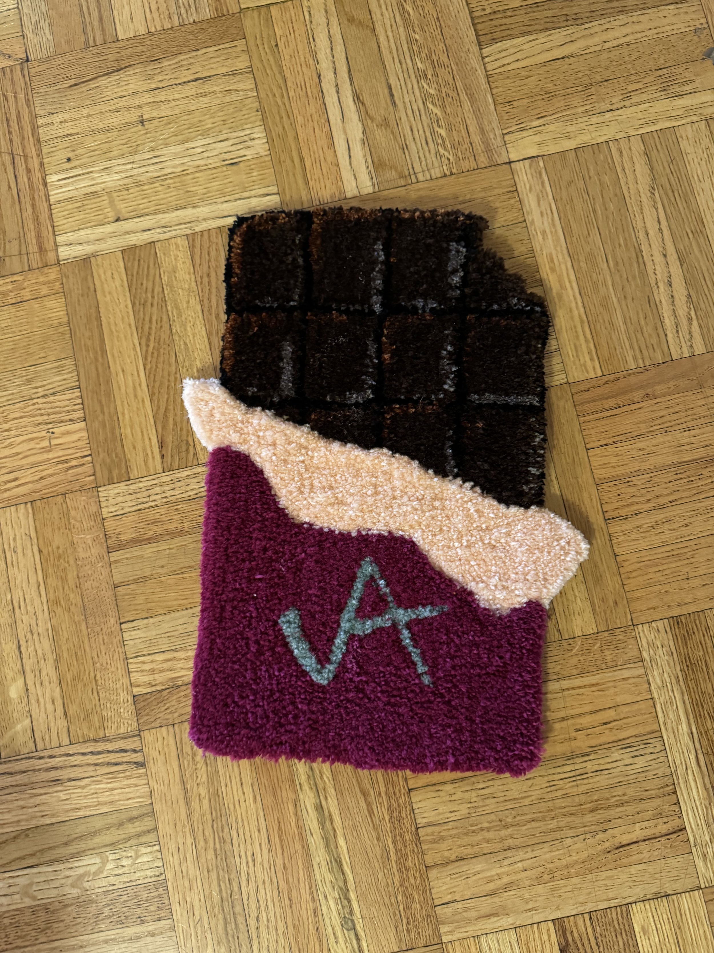 A small, rectangular multicolored carpet or rug with geometric pattern, placed on a wooden floor. The carpet has a pink, cream, and dark brown section, with a gray symbol or signature on the pink section.
