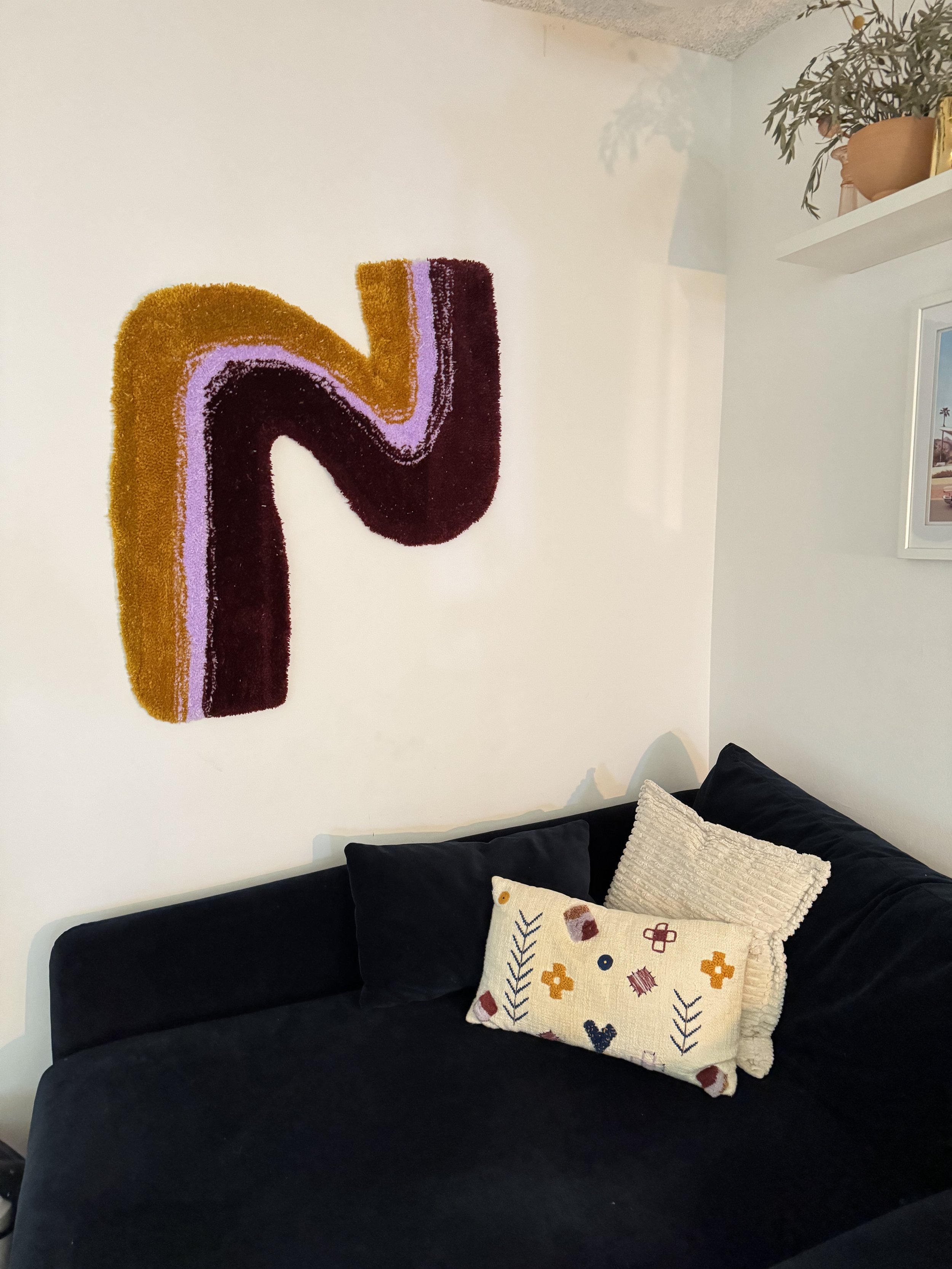 Living room corner with a black couch, decorative pillows, a colorful abstract wall hanging, and a white shelf with a potted plant.