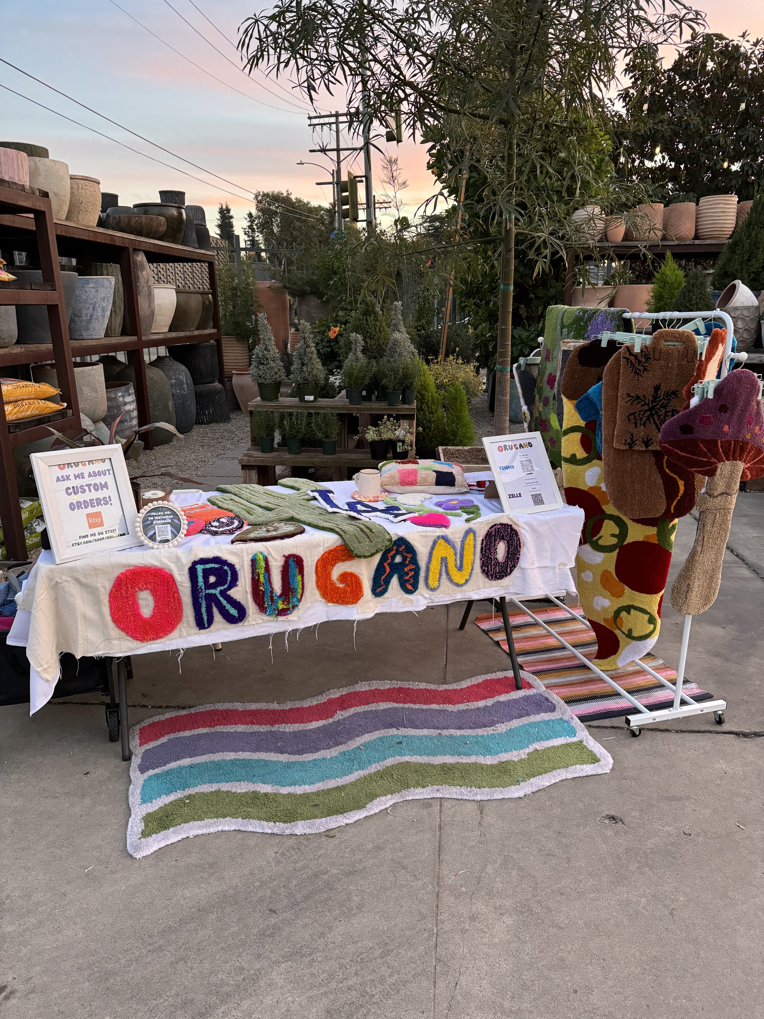 Outdoor craft market stall selling handmade colorful yarn items, including letters spelling 'ORUGANO,' with potted plants and garden supplies in the background at sunset.