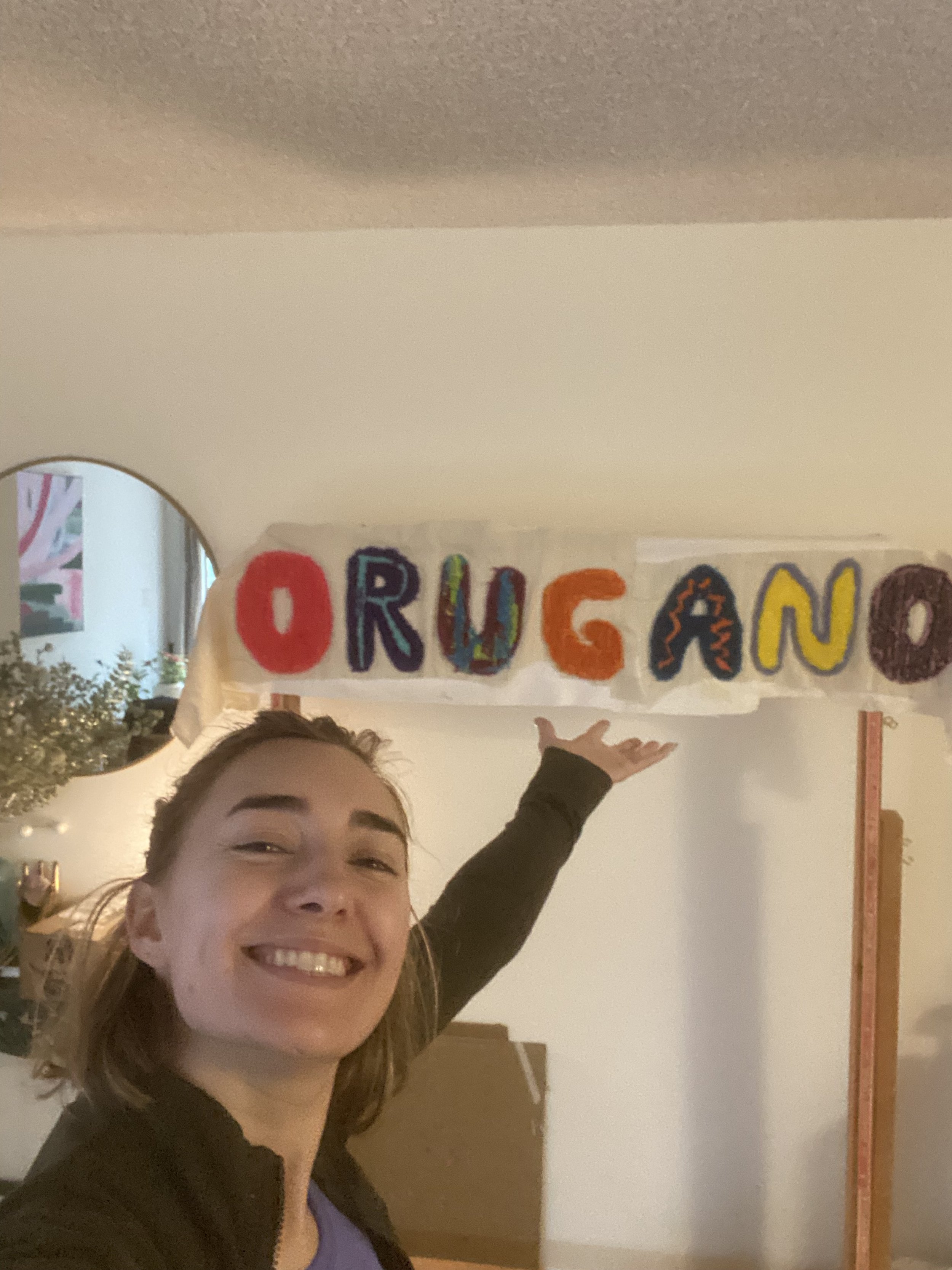 A smiling woman points to a colorful handmade sign that says 'ORUGANO' with each letter in a different color and style, indoors with a mirror and a plant visible in the background.
