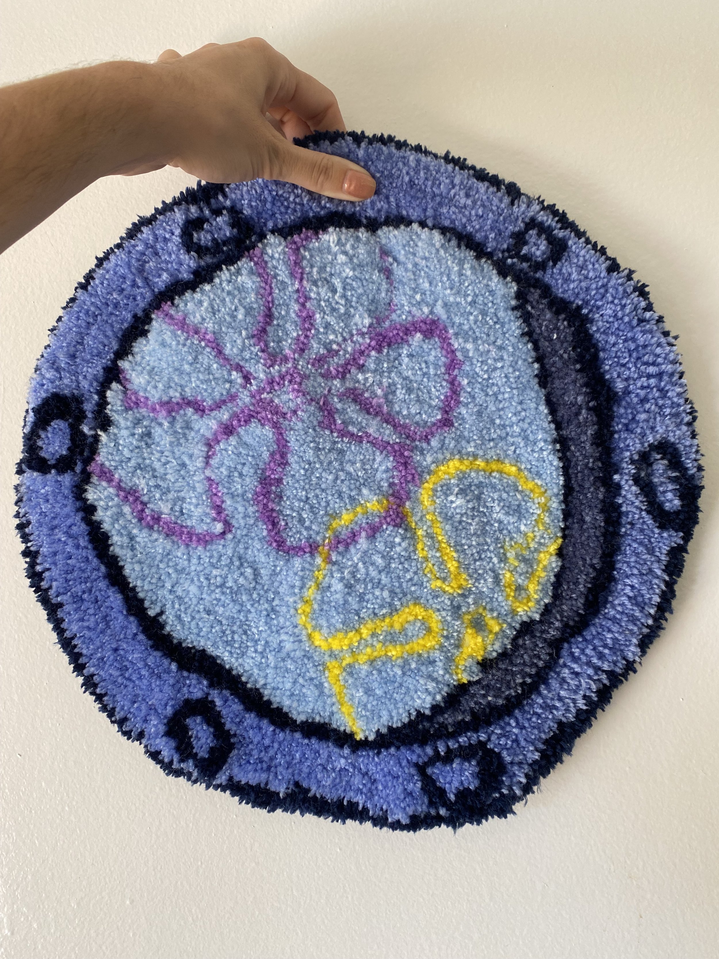 Close-up of a round, colorful carpet with a design of a purple butterfly and a yellow butterfly on a light blue background, held up by a person's hand against a white wall.