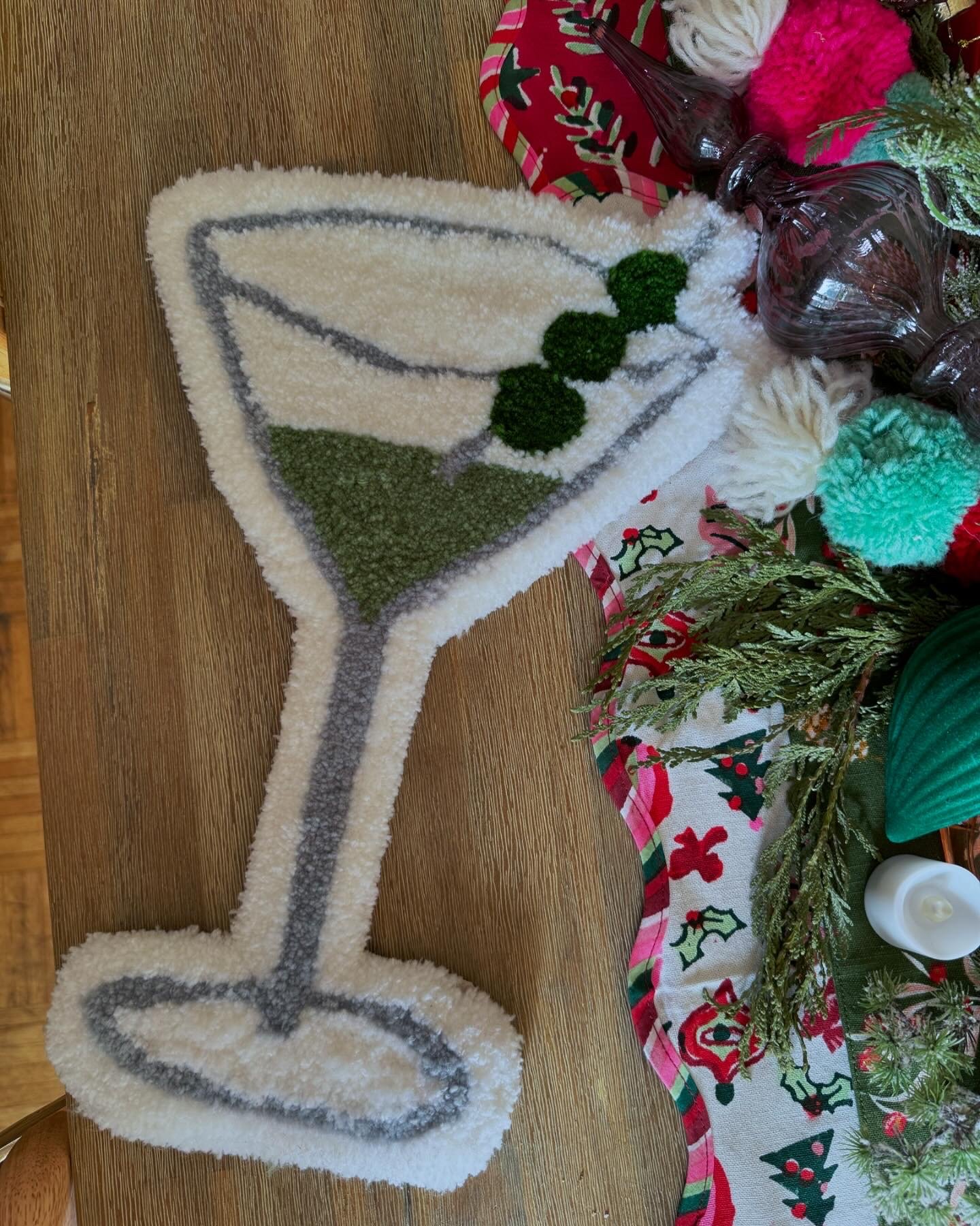 A decorative Christmas rug in the shape of a wine glass with a green olive skewer on top, placed on a wooden table amidst holiday decorations.