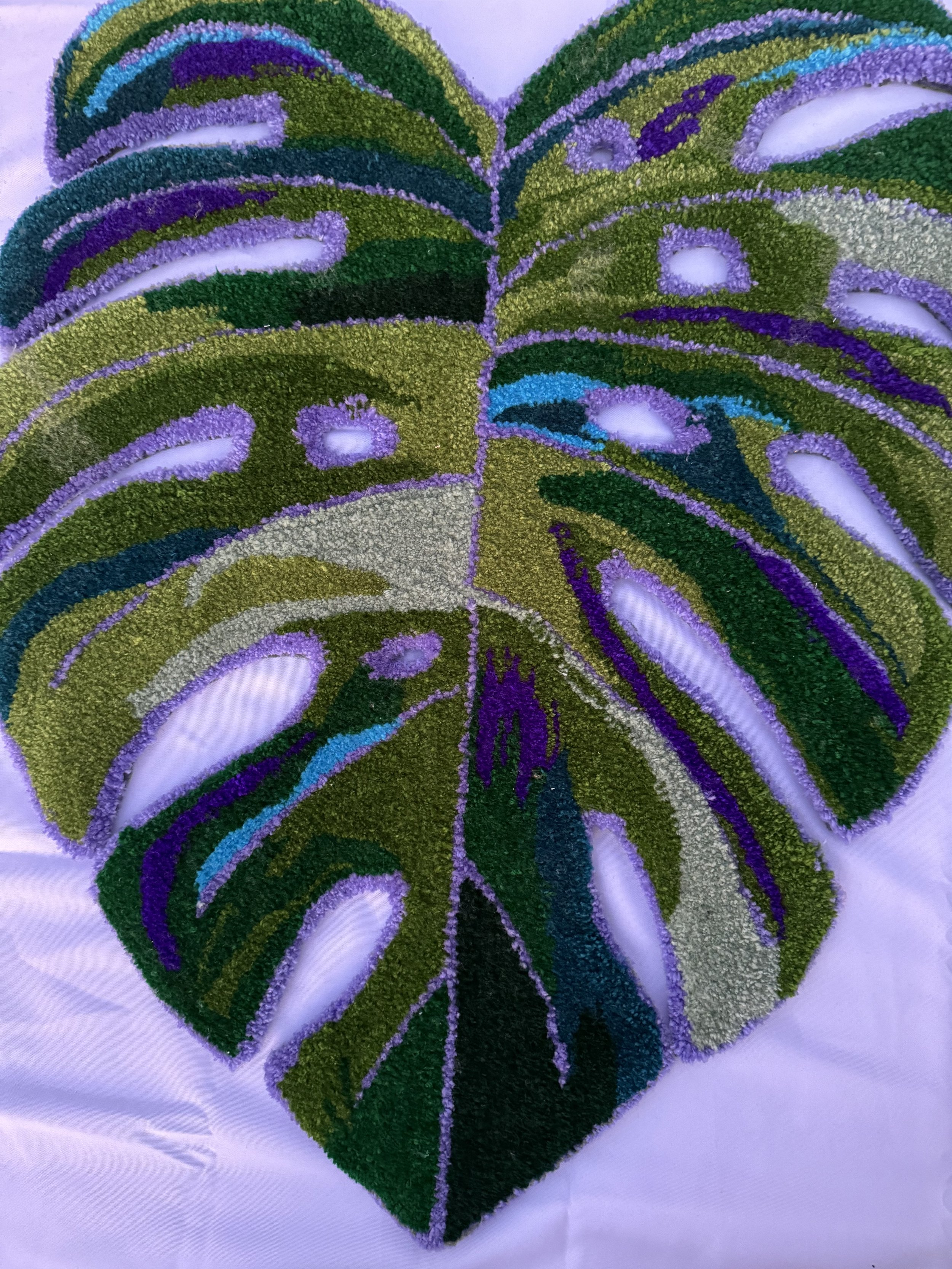 Colorful art piece of a large Monstera leaf made with colored sand or small beads, with shades of green, purple, blue, and white, laid on a white surface.