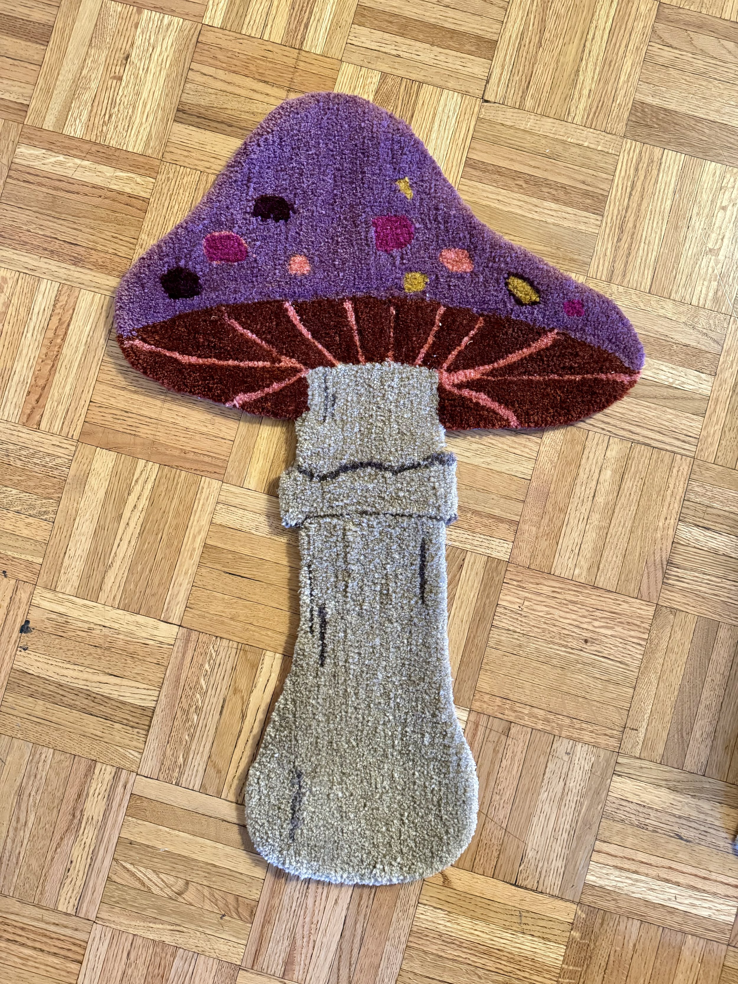 A colorful rug shaped like a mushroom with a purple cap decorated with multicolored spots and a beige stem, placed on a wood floor.