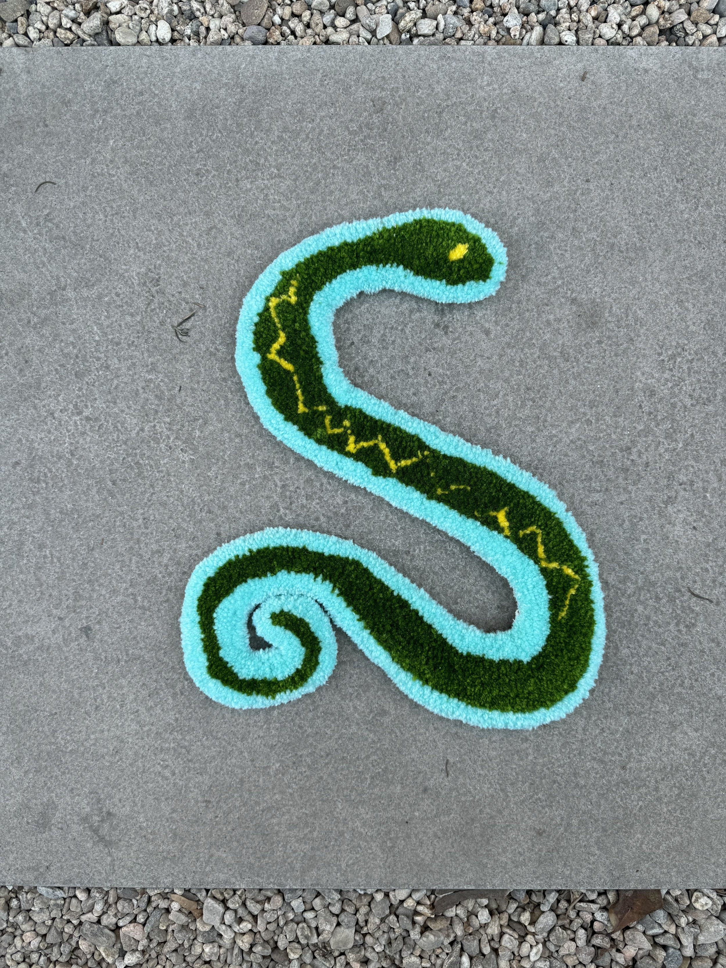 Colorful decorative rug of a snake in the shape of a letter S on a concrete surface with gravel around.