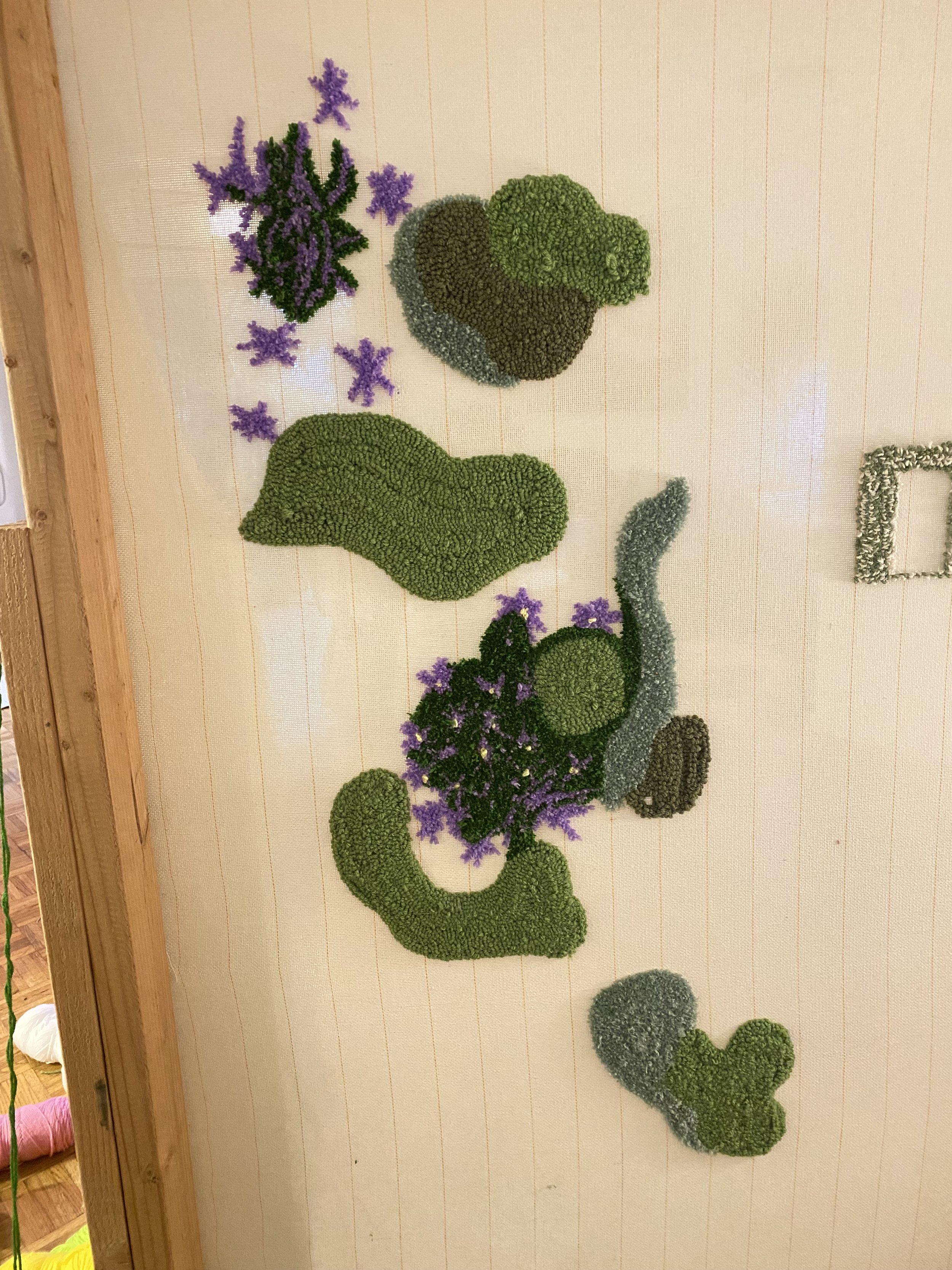 A vertical wall hanging made of various colored moss and artificial flowers arranged in abstract shapes on a beige wall.