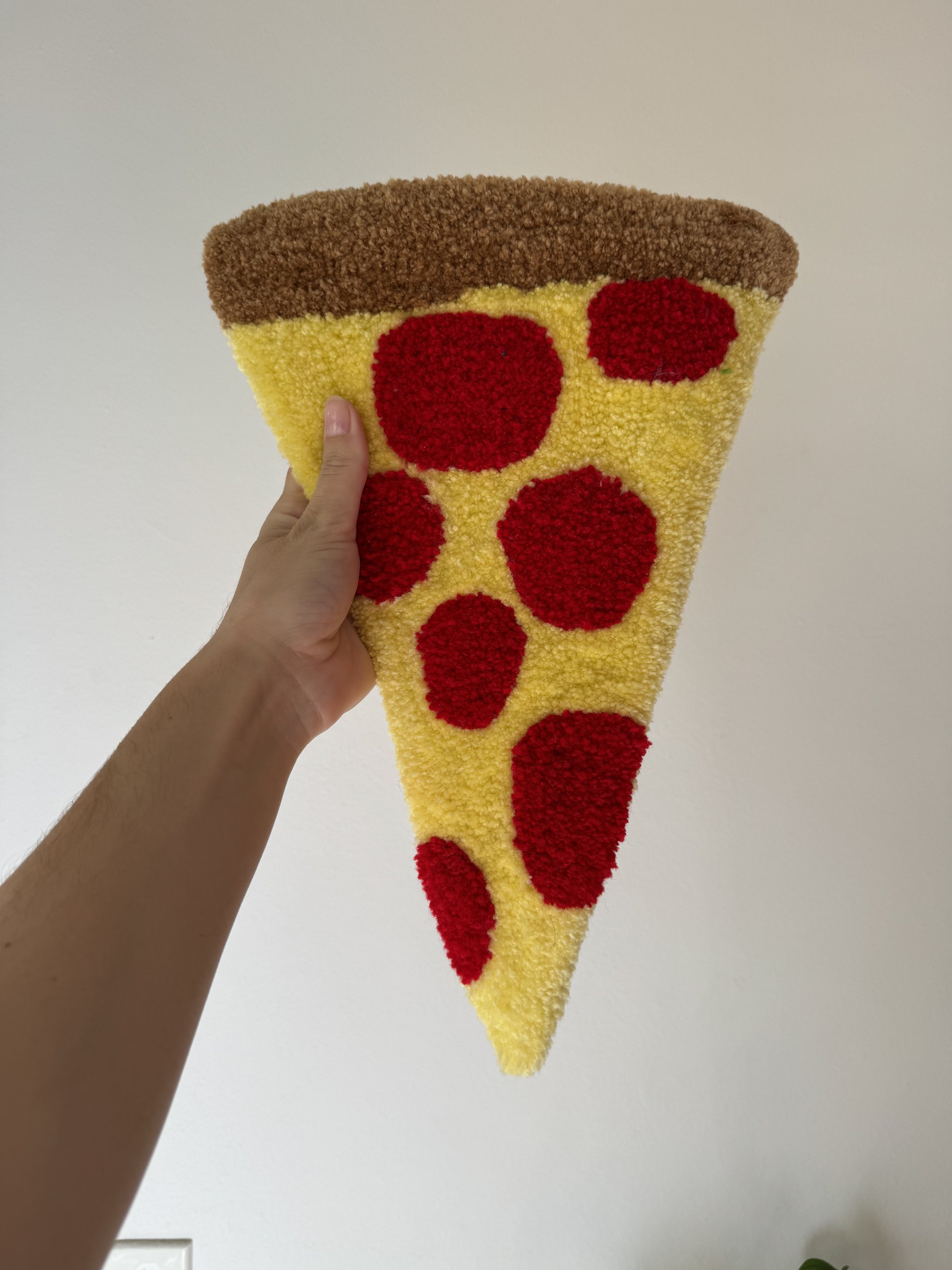 A person holding a fuzzy, carpet-like rug shaped like a slice of pepperoni pizza with a brown crust, yellow cheese, and red pepperoni toppings against a plain white wall background.