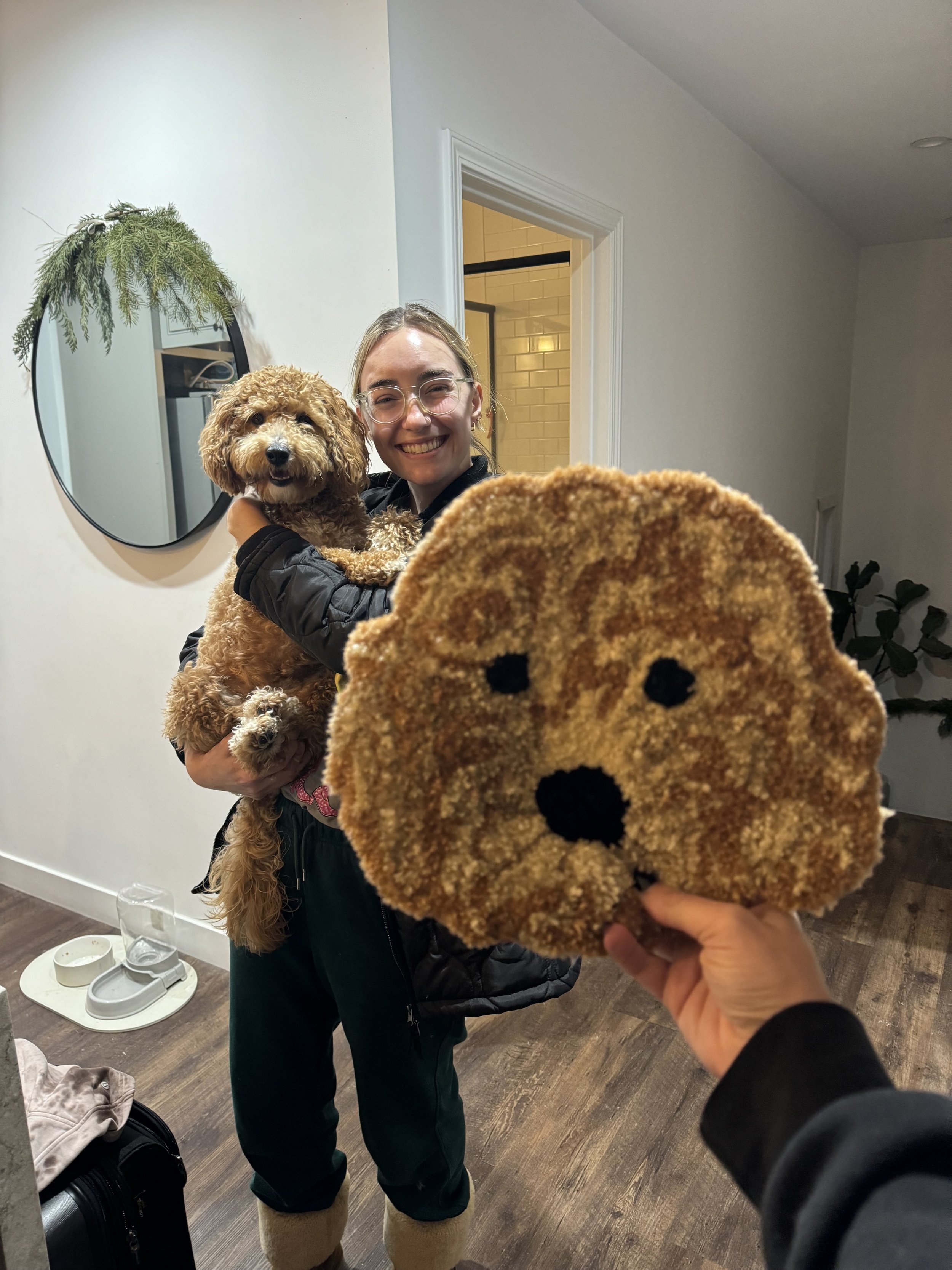 A woman is holding a small, curly-haired dog and smiling at the camera. Someone in the foreground is holding a dog-shaped carpet or rug, also facing the camera. The scene is indoors with white walls and wooden floors.