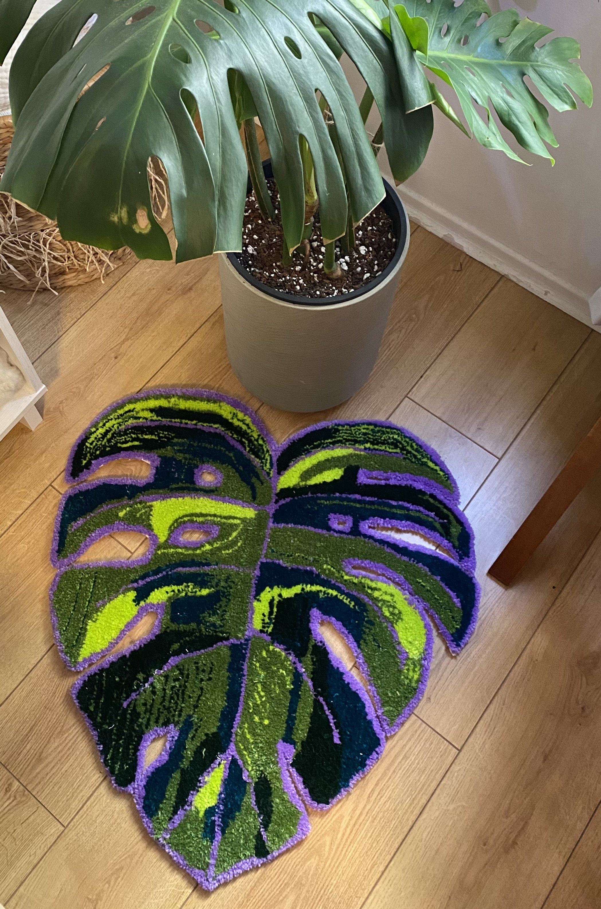 A large green monstera leaf-shaped doormat on a wooden floor, with a potted monstera plant nearby.