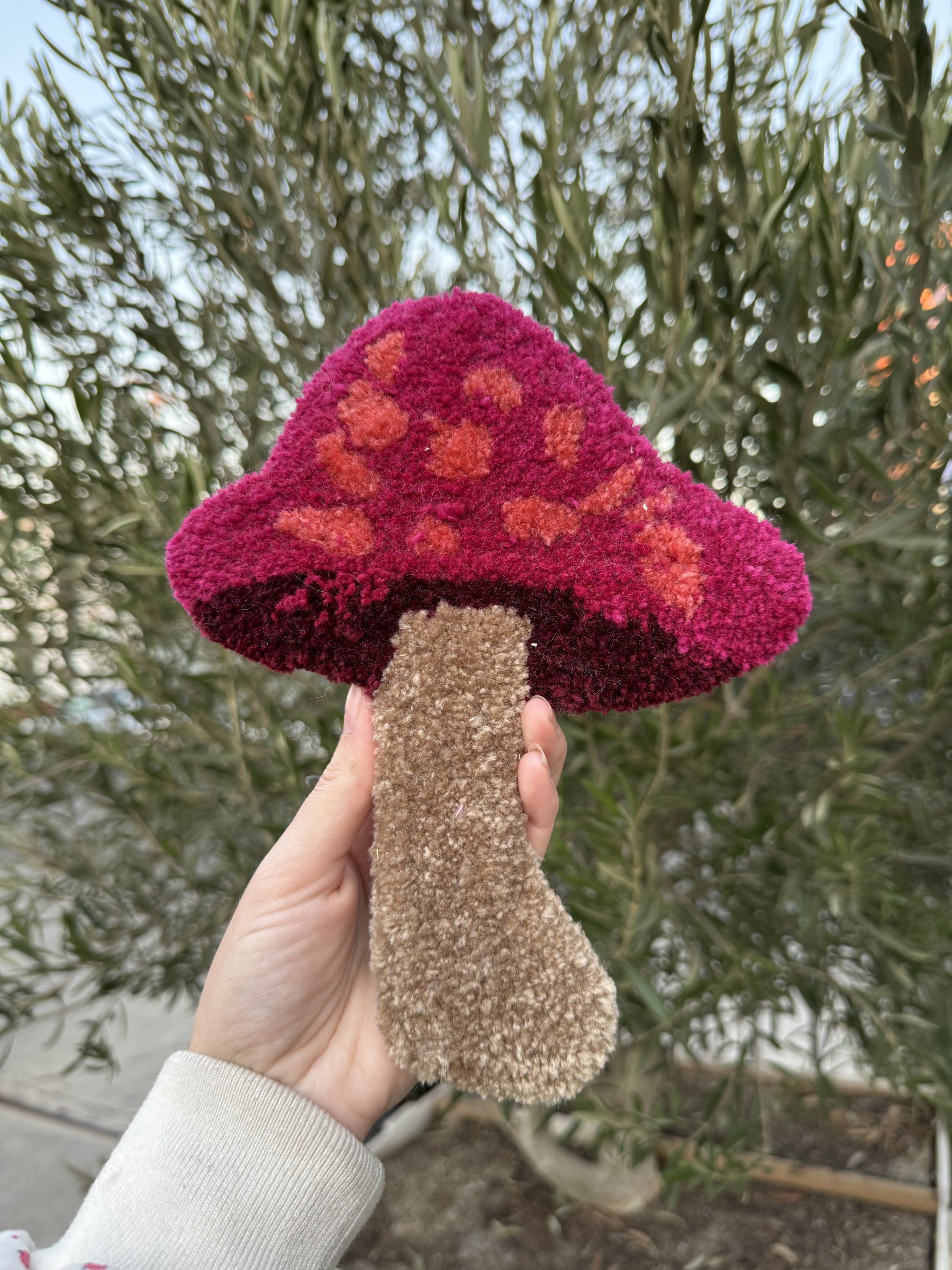 Hand holding a plush mushroom-shaped toy with a red cap decorated with orange spots and a brown stem, outdoors with green bushes in the background.
