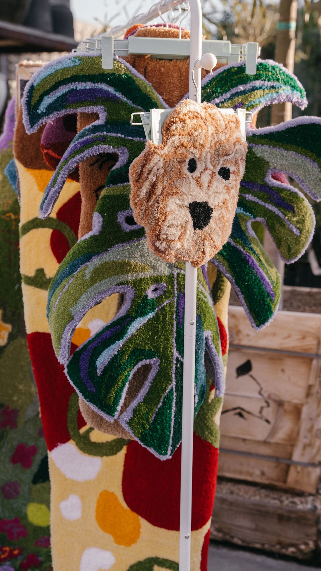 Colorful floral-themed rugs hanging on display at an outdoor market. One rug features a large, ornate green tropical leaf pattern, and another has various bright, abstract shapes. A small, plush dog face headband is hanging in the foreground.