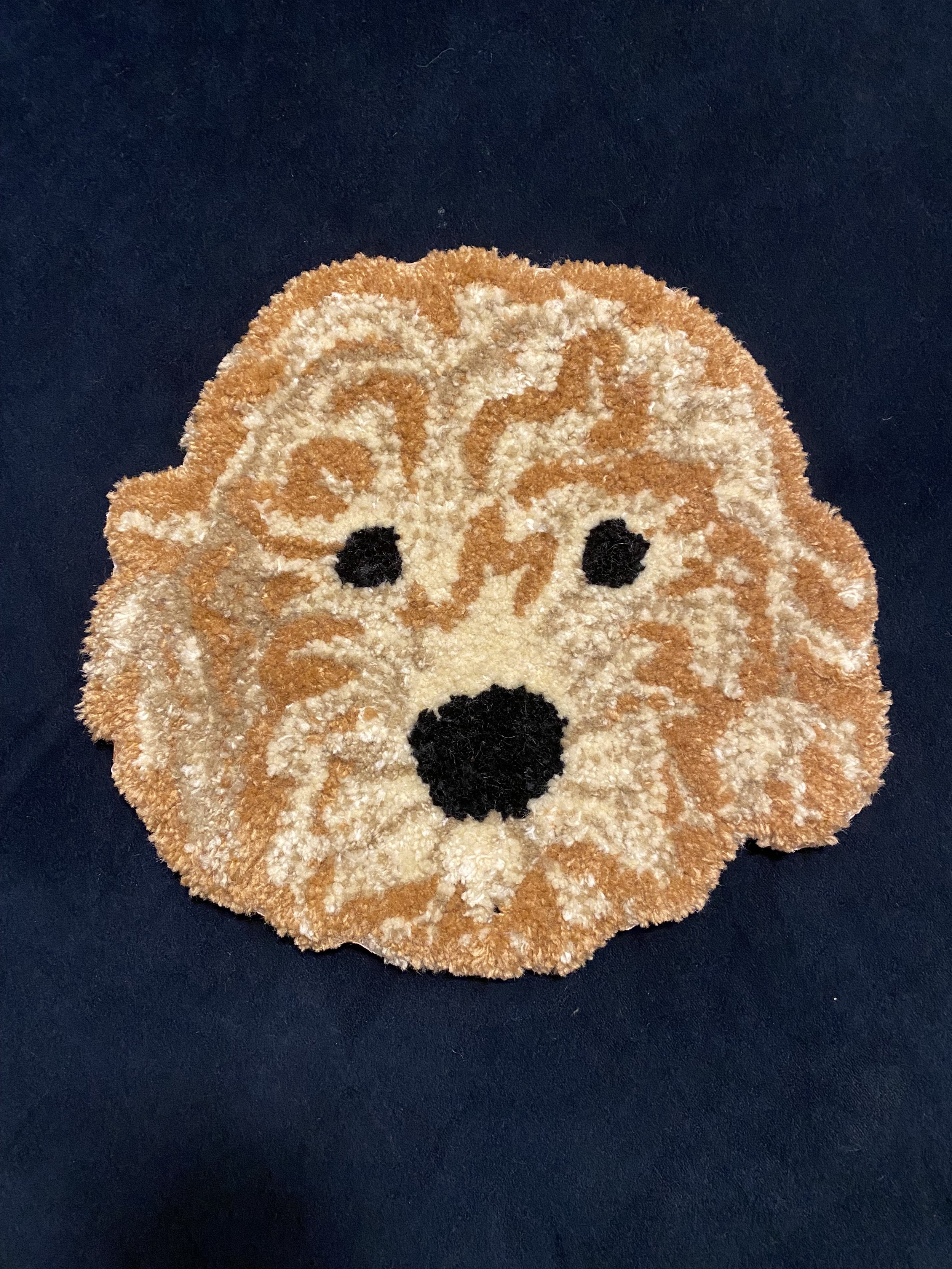A small, round, plush rug with a design of a dog's face, featuring black eyes, a black nose, and light brown and cream detailing, placed on a dark surface.