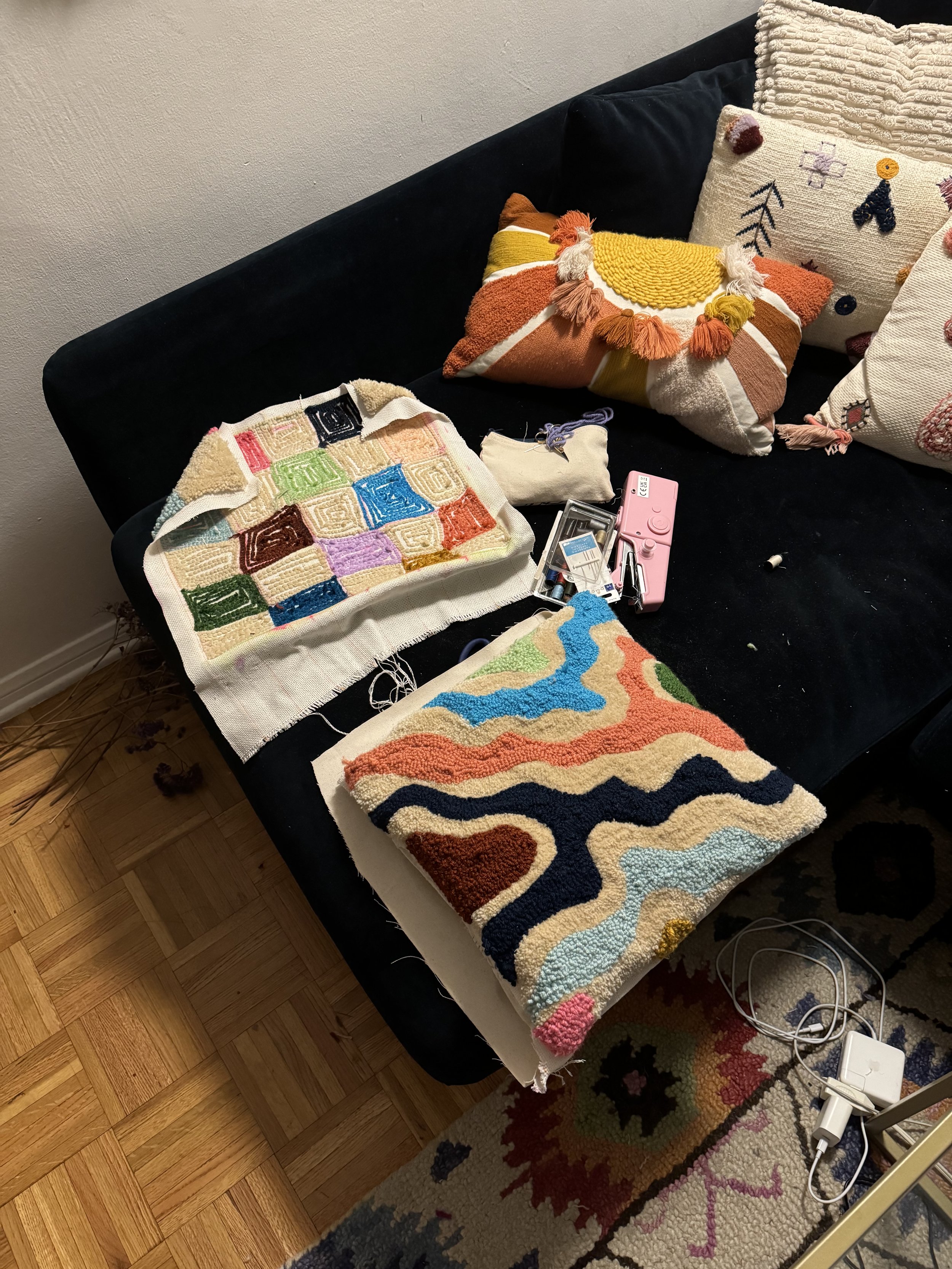 A black sofa with assorted patterned and textured pillows, unfinished colorful tapestry art projects, a pink needle felting kit, and various crafting supplies on the table.