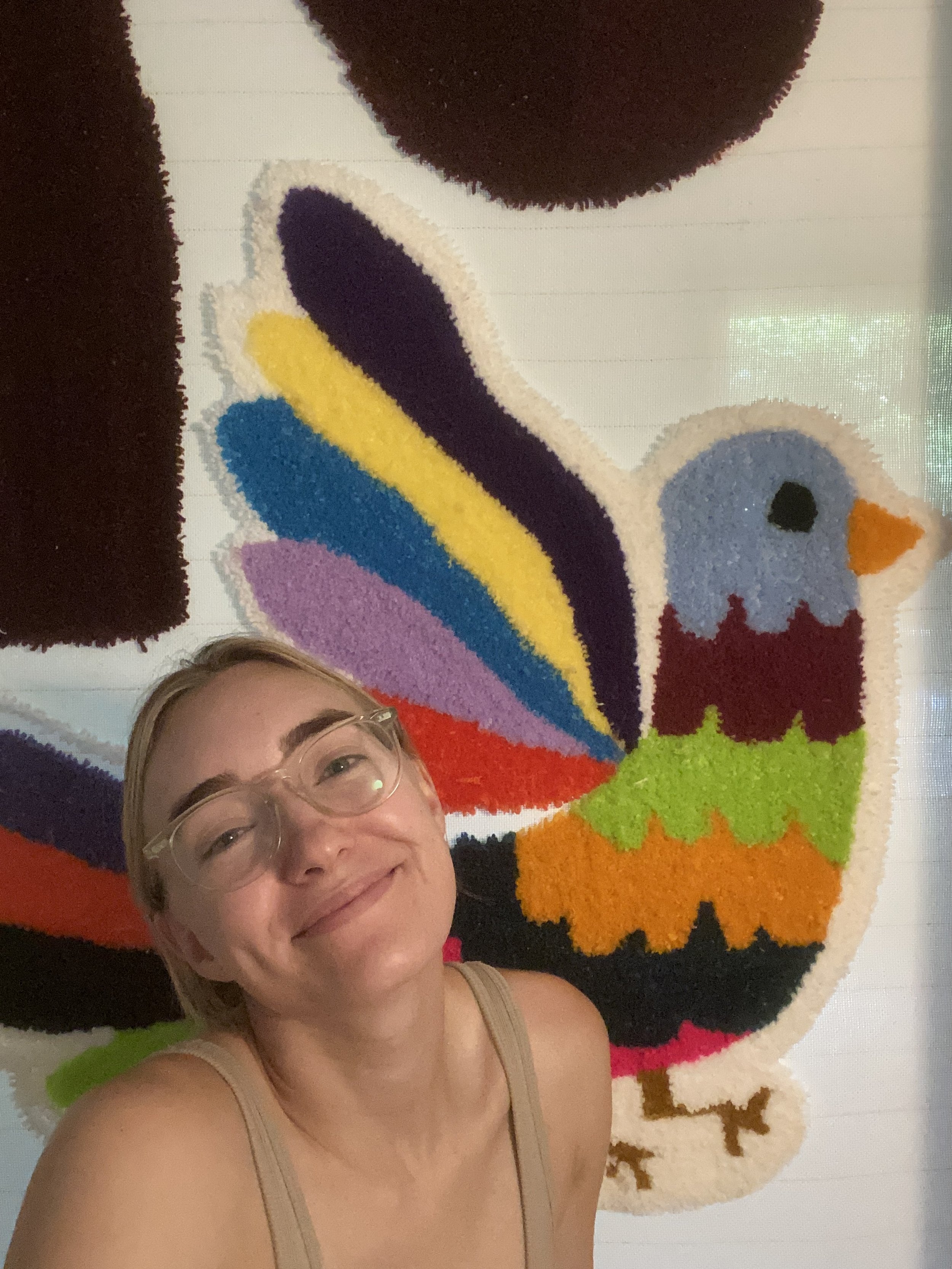 A smiling woman with glasses taking a selfie in front of a colorful, cartoonish rooster rug on the wall.