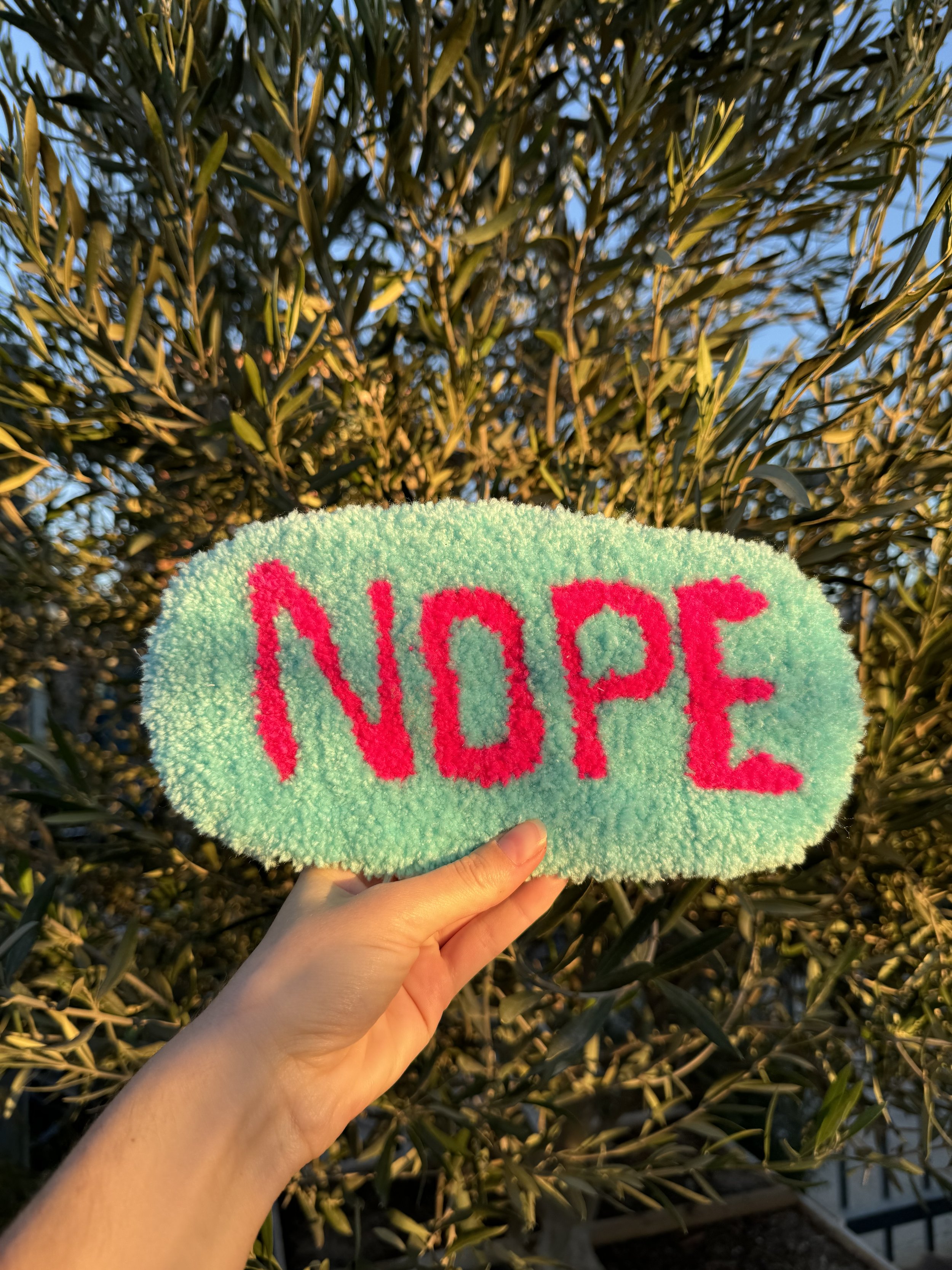 A hand holds a fluffy, oval-shaped item with the word 'NOPE' written in pink on a light green background, with a leafy plant in the background.