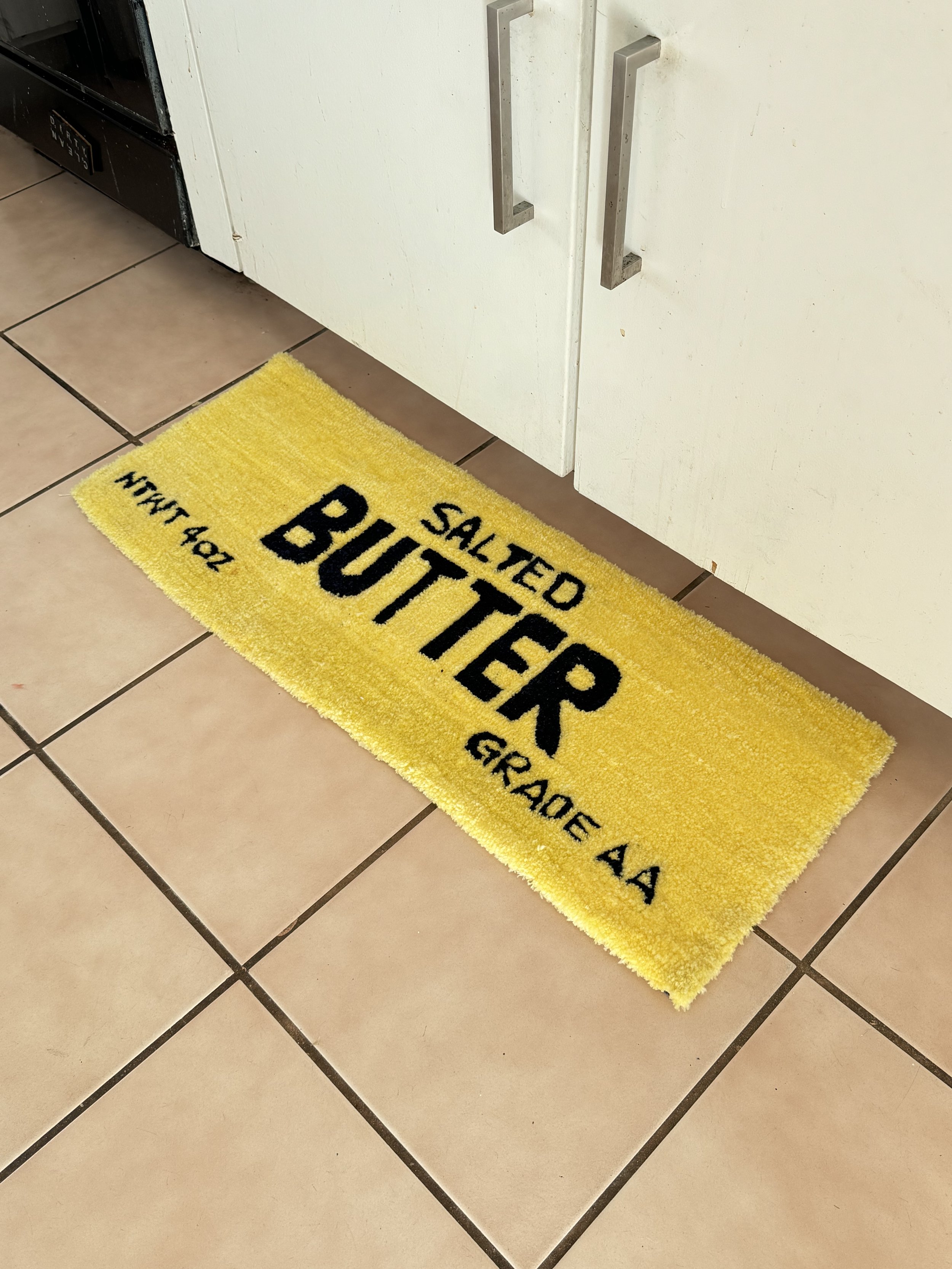 A yellow bath mat with black text that reads 'Salted Butter' in a kitchen, placed on a tiled floor near white cabinets.