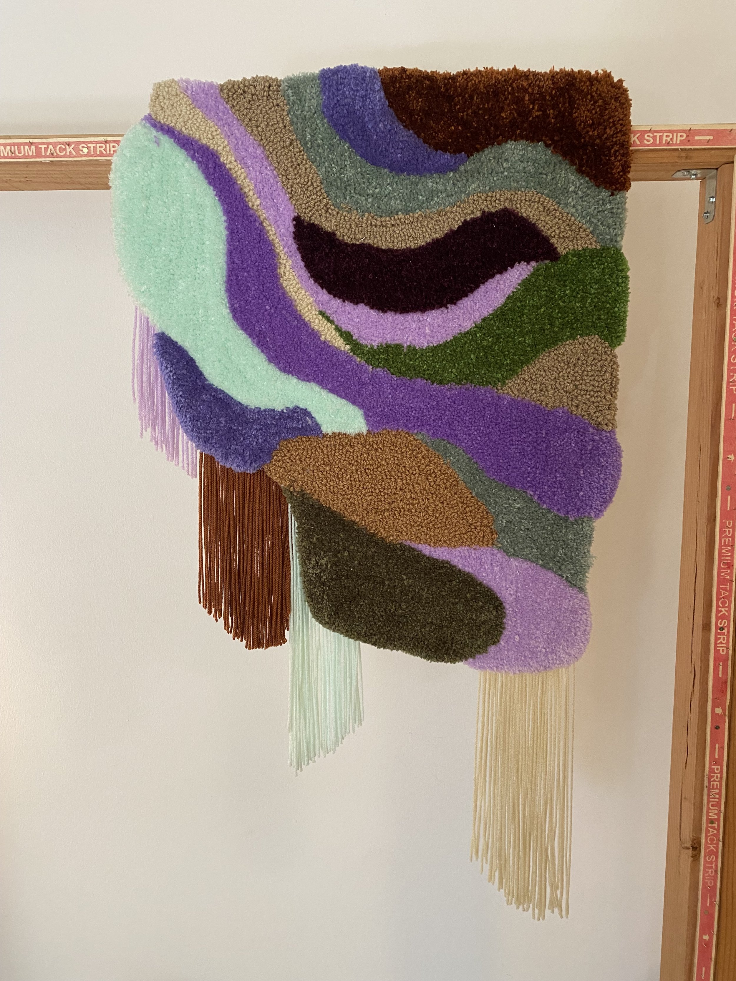 Colorful abstract textile wall hanging with wavy patterns and fringes at the bottom, mounted on a wooden frame.
