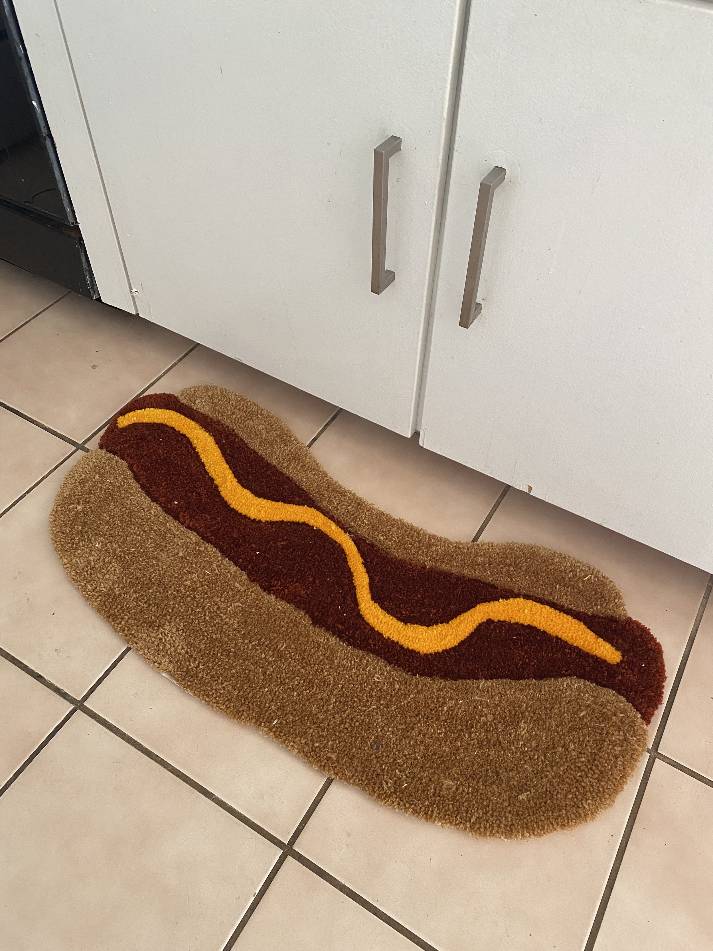 A decorative rug with a wavy pattern in brown, yellow, and orange is placed in front of white kitchen cabinets with silver handles, on beige tiled flooring.