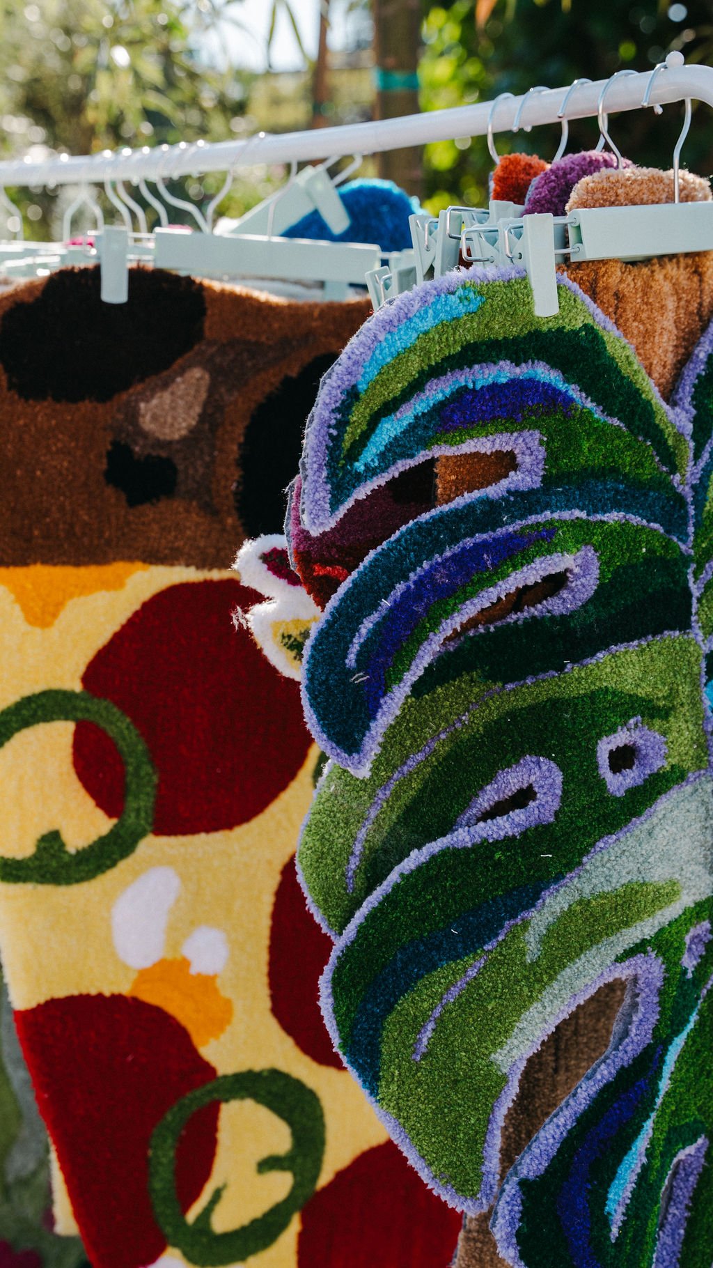 Colorful patterned rugs hanging outdoors on a clothesline, with a background of green trees and bright sunlight.