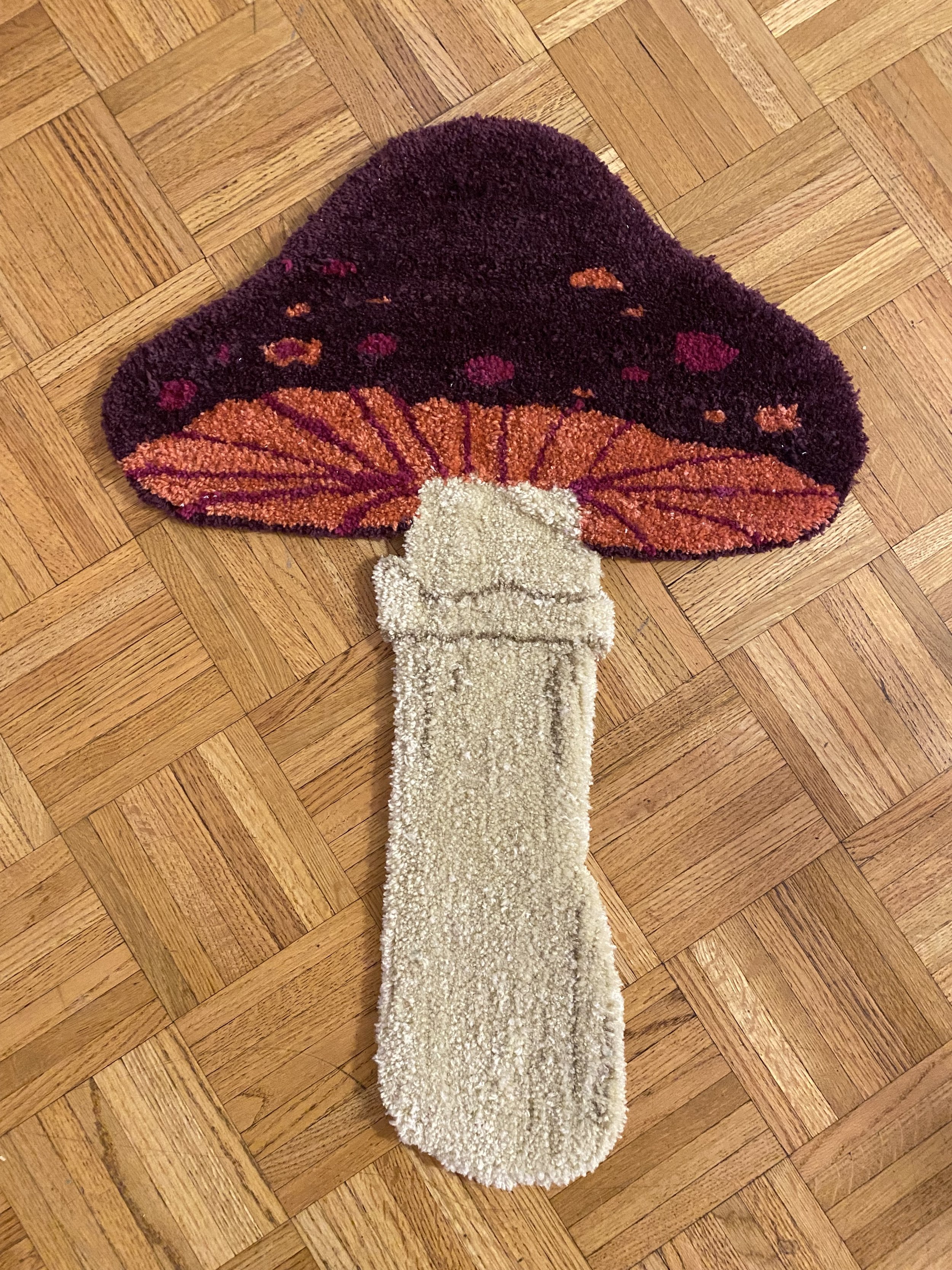 A rug designed to look like a mushroom, with a beige stem and a multicolored cap in shades of purple, orange, and red, placed on a wooden parquet floor.