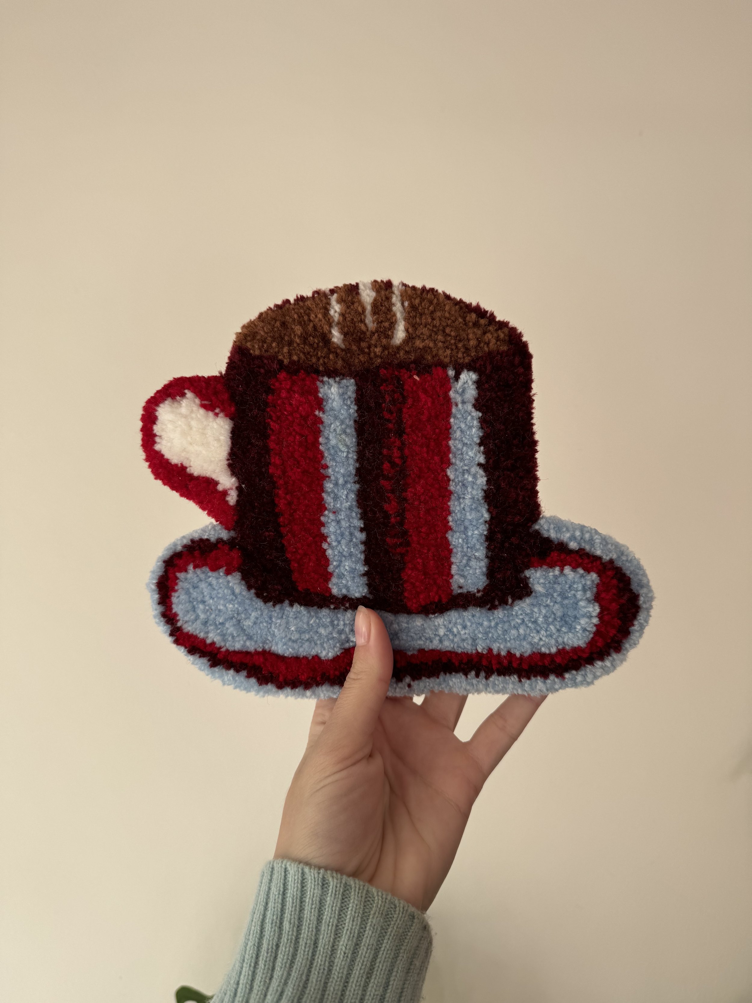 A hand holding a knitted mug and saucer, designed to look like a coffee mug with a handle, with red, brown, white, and light blue colors.