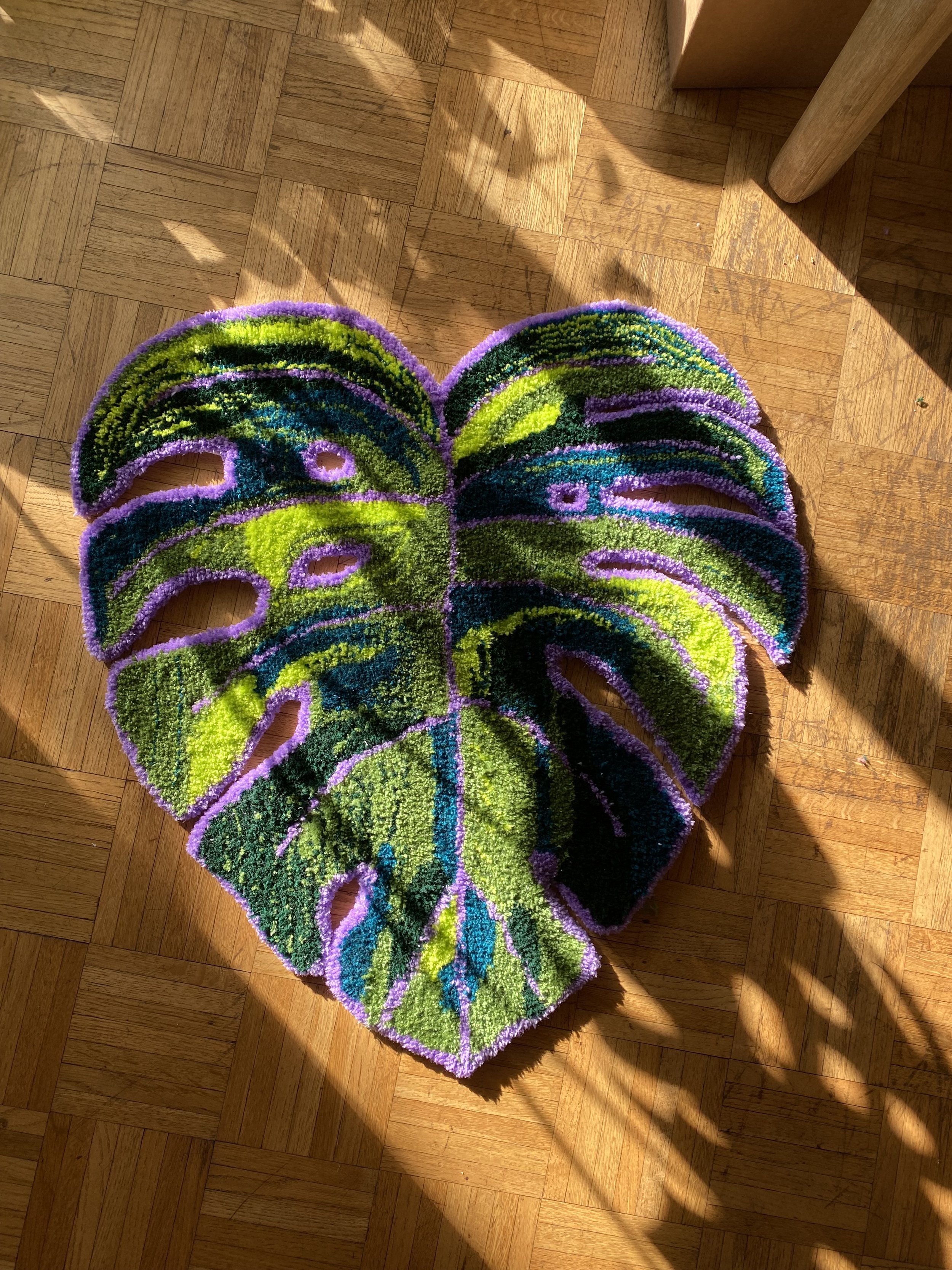 Colorful monstera leaf-shaped rug with purple border on hardwood floor, sunlight and shadows.