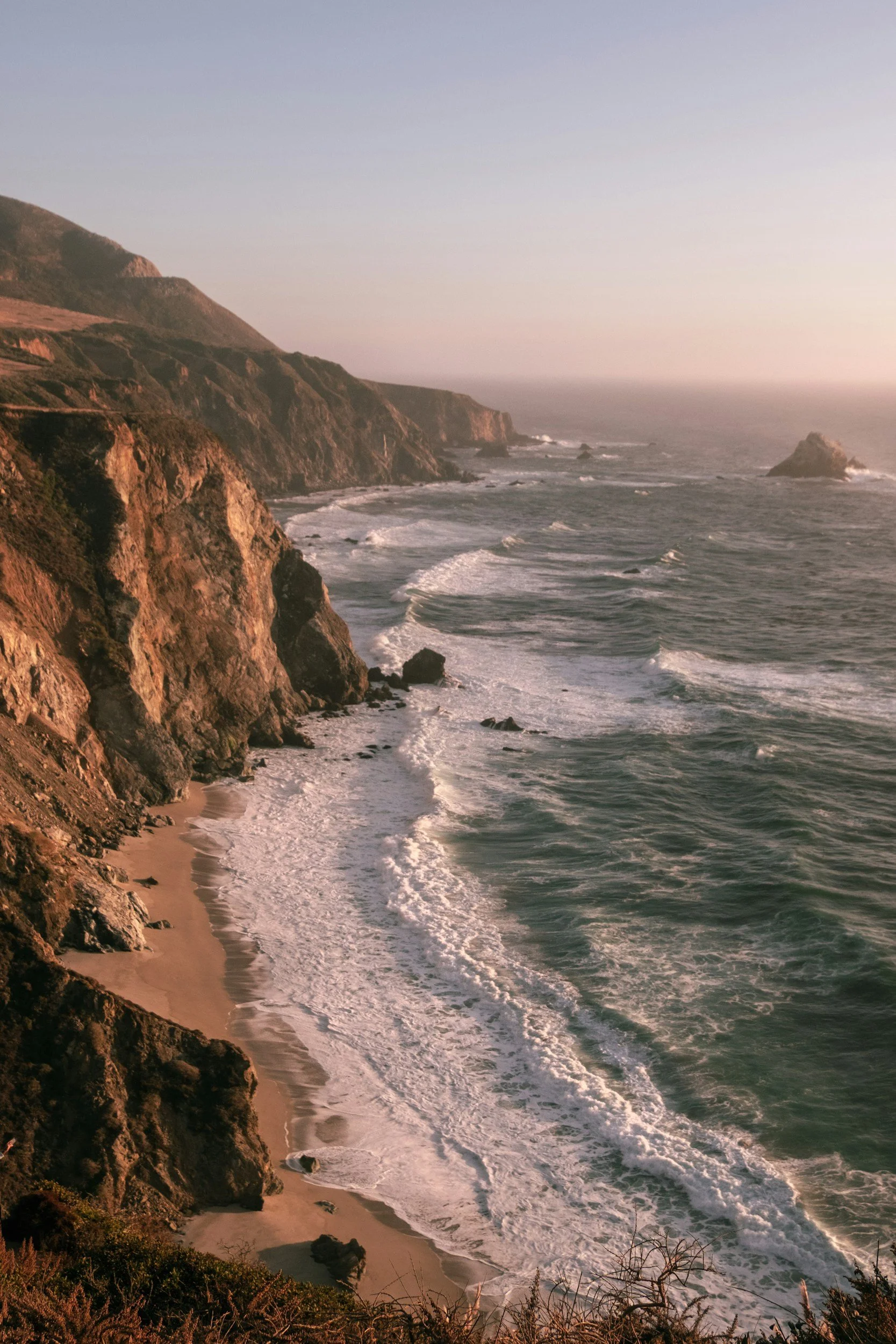 Image of the gorgeous Big Sur coastline with waves gently coming into shore and a dramatic cliff, taken around sunset with a warm haze in the seaspray, invoking the importance of centering the land and our relationship with it in astrocartography.