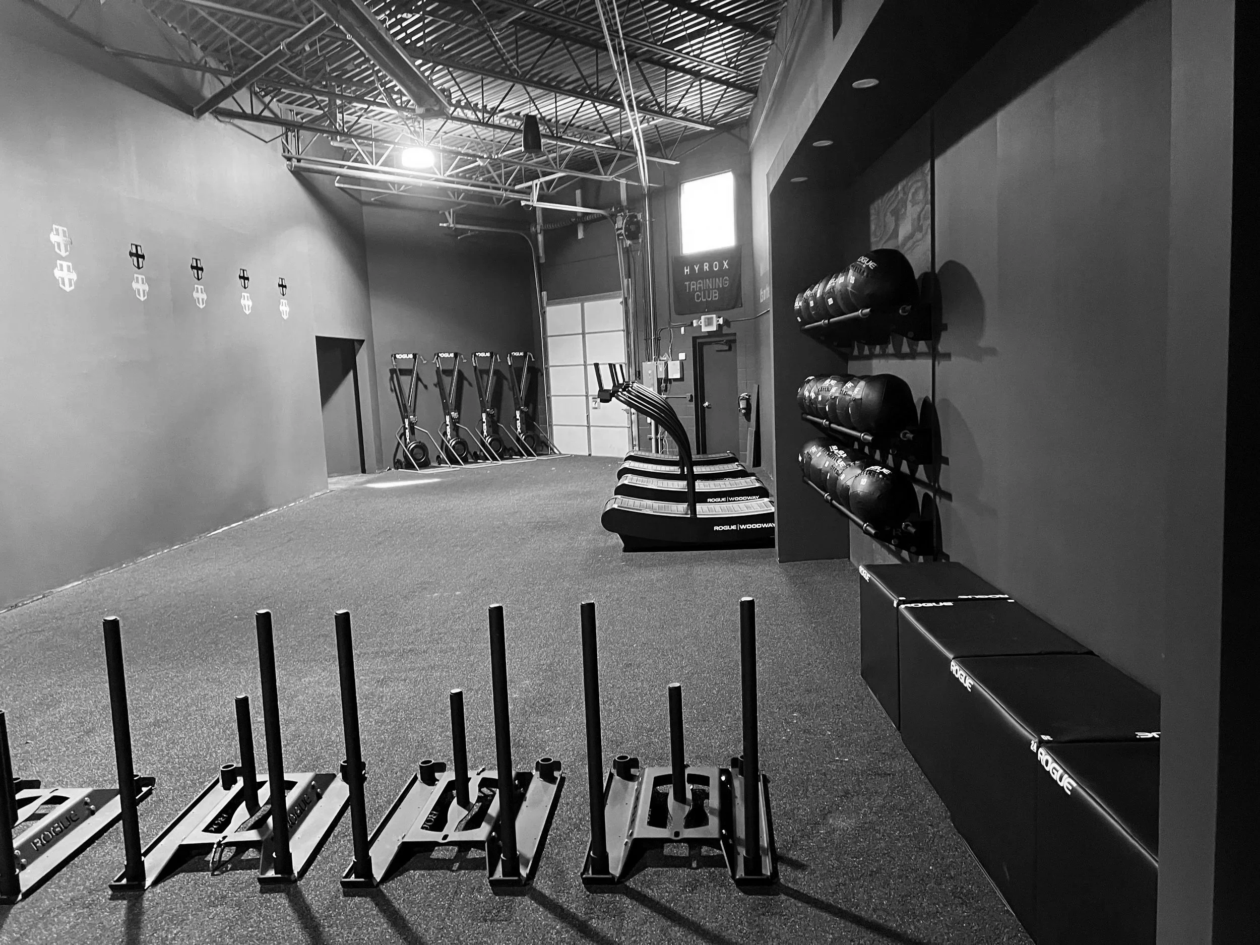 Empty gym with black and white color scheme, featuring wall-mounted medicine balls, stacked on shelves, staggered plyometric boxes, and gymnastic rings hanging from the ceiling, with a motivational sign that reads 'HYROX TRAINING CLUB' and a door in the background.