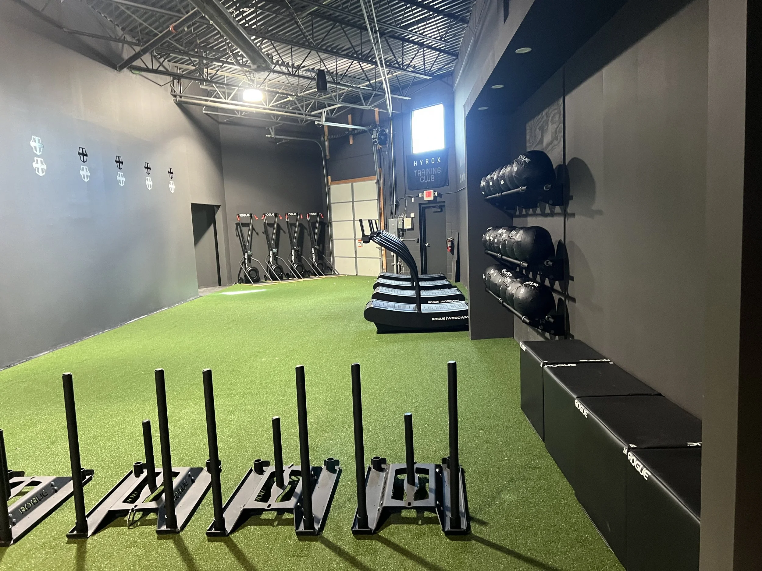 Indoor gym with black walls, green turf flooring, and exercise equipment including sleds, medicine balls, and benches, under a high ceiling with metal support beams.