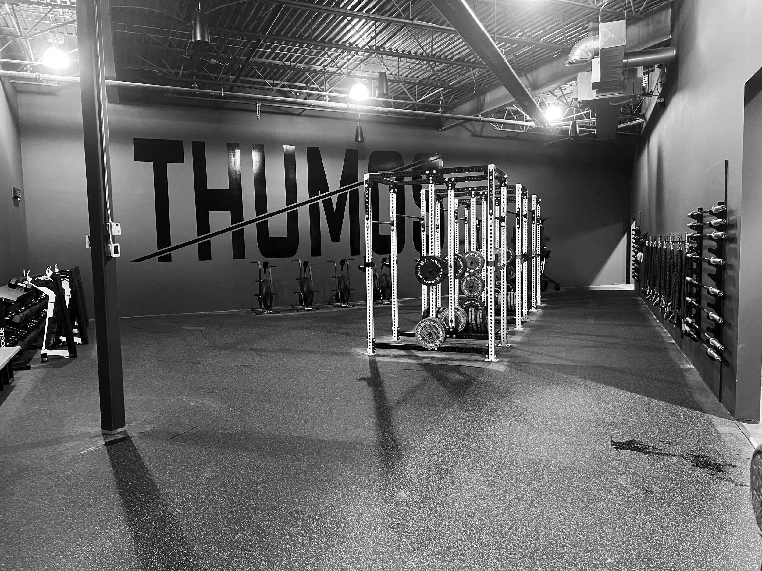 An empty gym with workout equipment including weight plates, Squat racks, and wall-mounted bars, with a large 'HIIT' sign on the wall and bright overhead lighting.