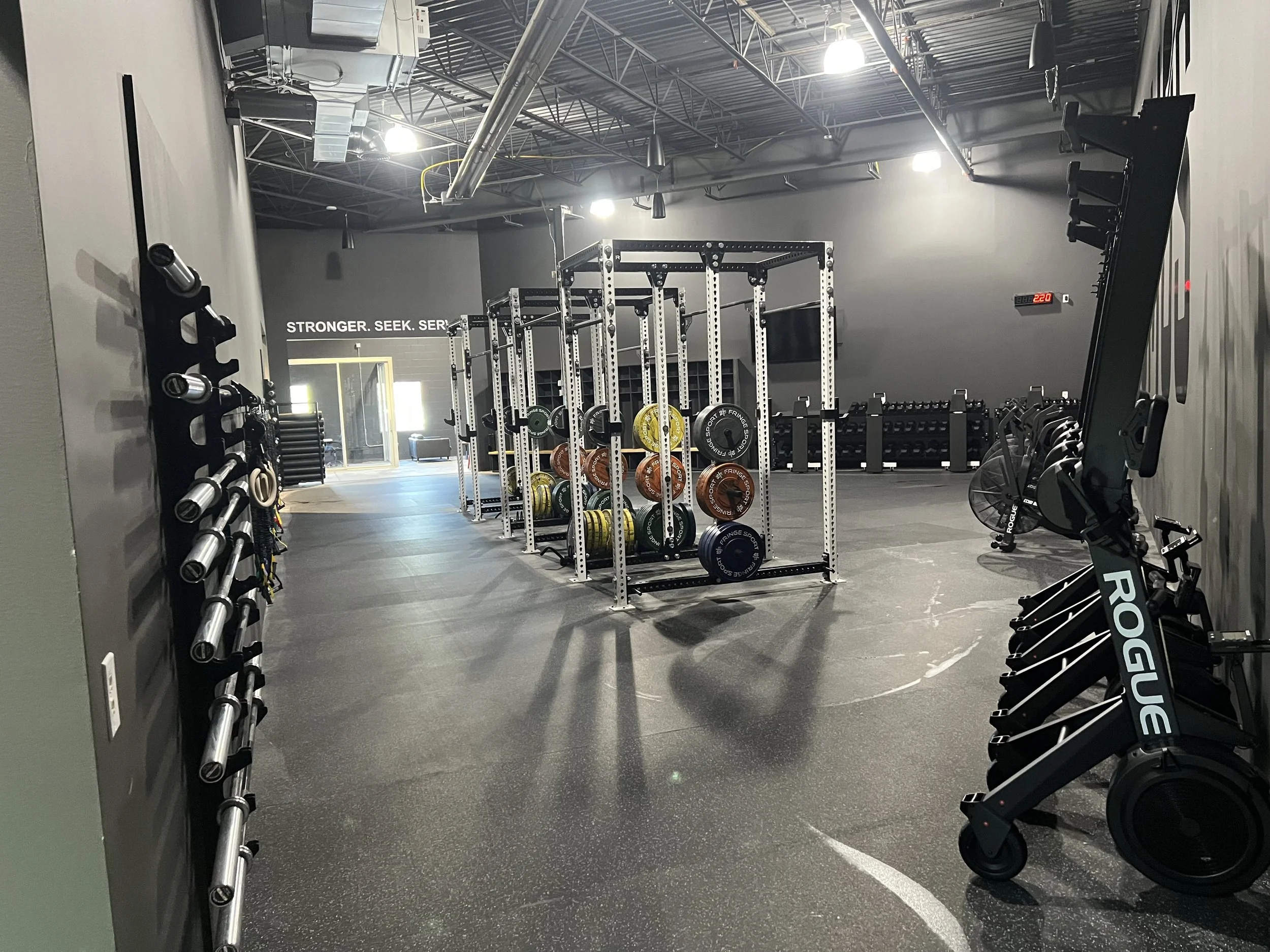 Gym with weightlifting equipment, squat rack with weight plates, dumbbells, and rowing machine, with motivational slogan 'Stronger. Seek. Serve.' on the wall.
