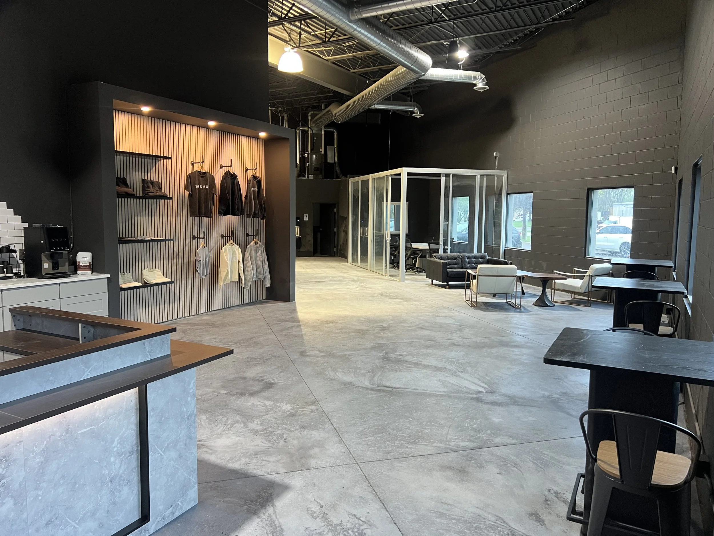 Modern commercial interior with black and gray walls, large windows, and a seating area with tables and chairs. There is a display wall with clothing and shoes, and a coffee station in the corner.