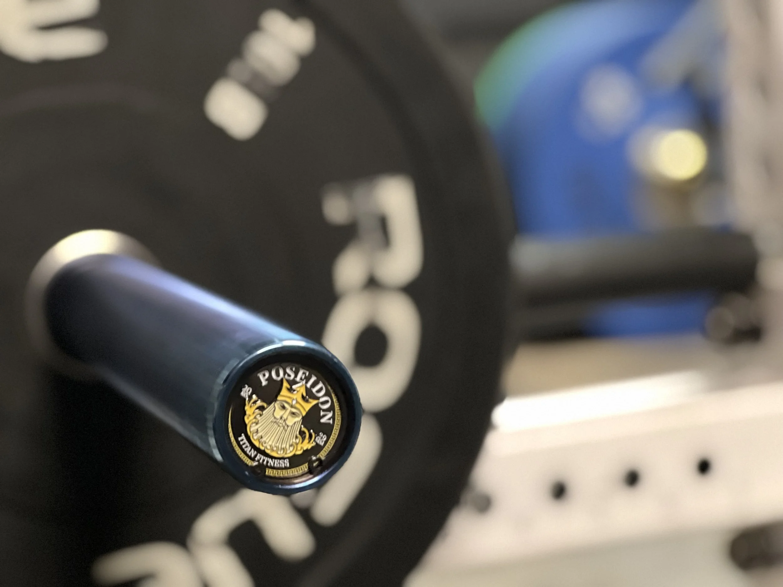 Close-up of a black dumbbell handle with a gold and black logo on its end cap, reading 'POSEIDON TRAIN FITNESS' with a stylized face and crown.